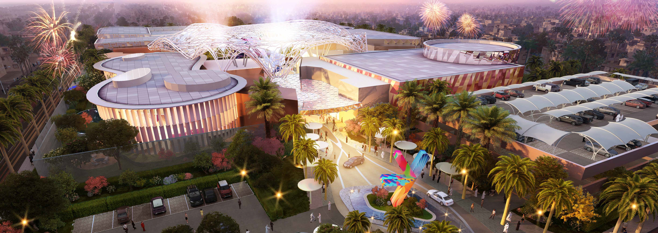 Aerial view of a modern entertainment complex with colorful structures, palm trees, and fireworks in the sky, with parking lots and people walking around.