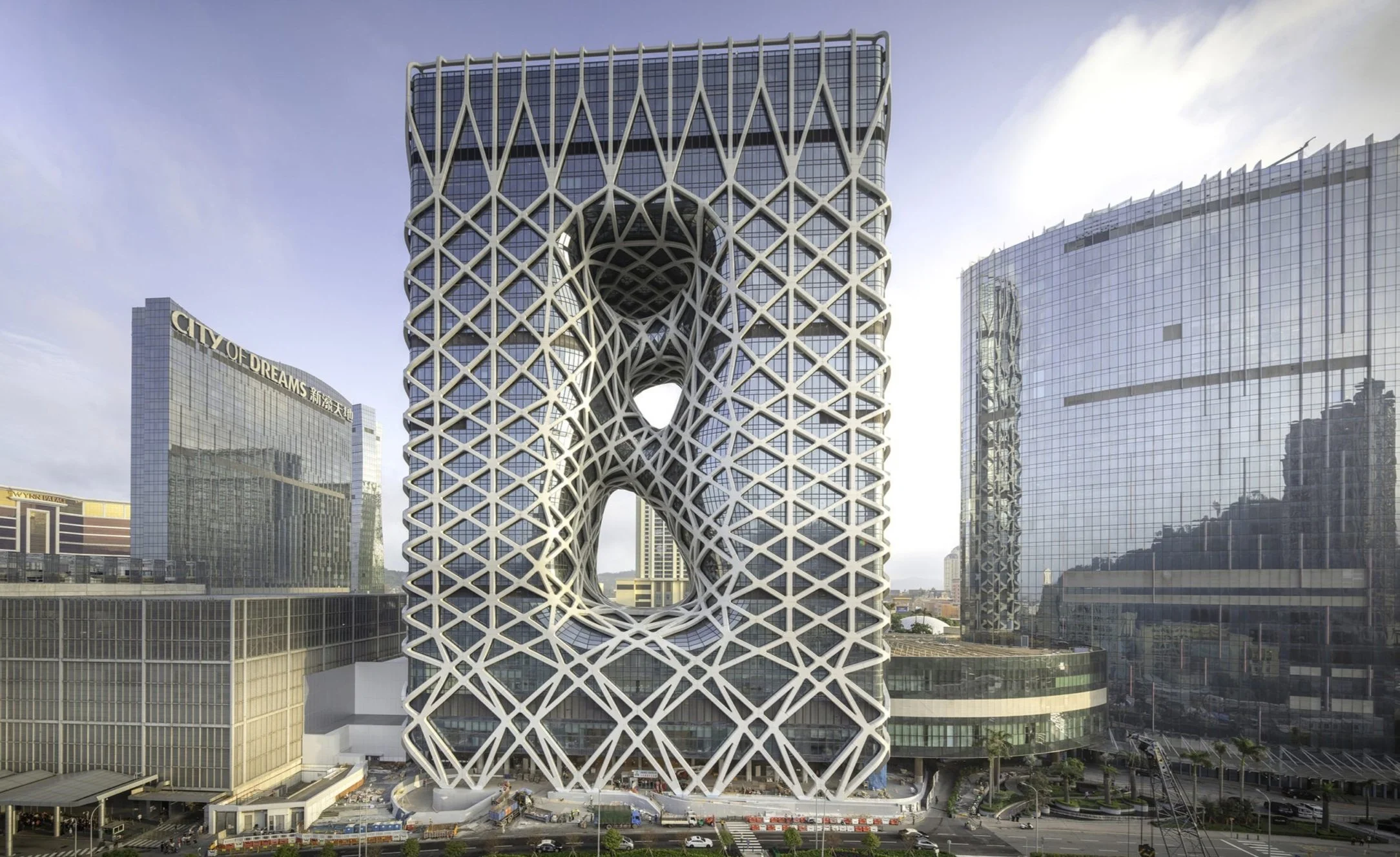 Futuristic skyscraper with a lattice-like exterior structure featuring three large circular openings, surrounded by other modern glass buildings in an urban cityscape.