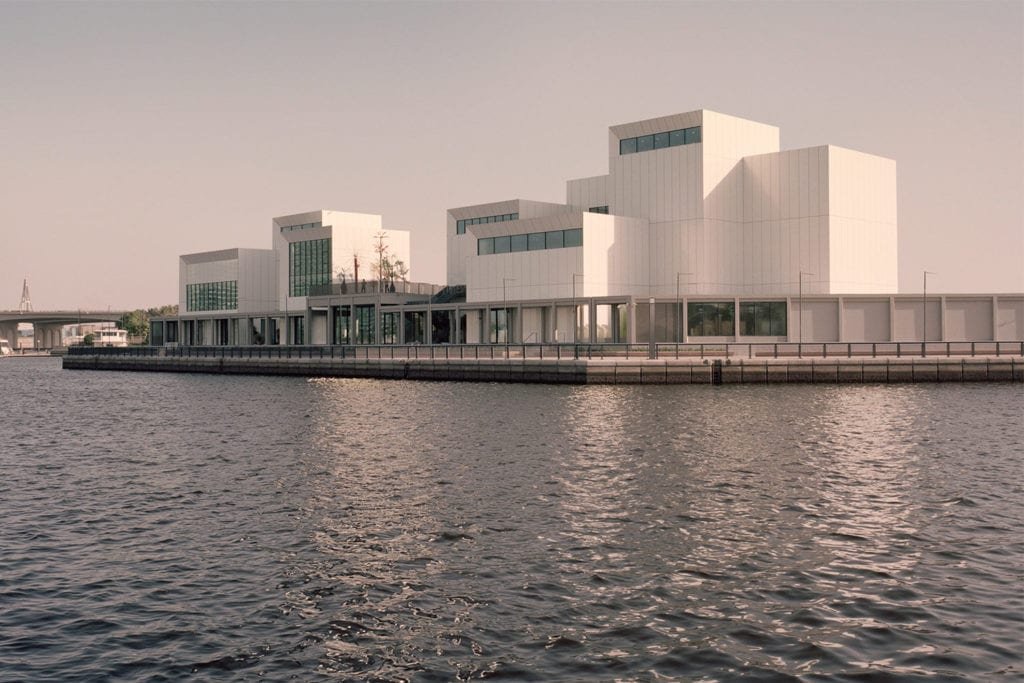 Modern white building with geometric shapes along a waterfront with water in the foreground.