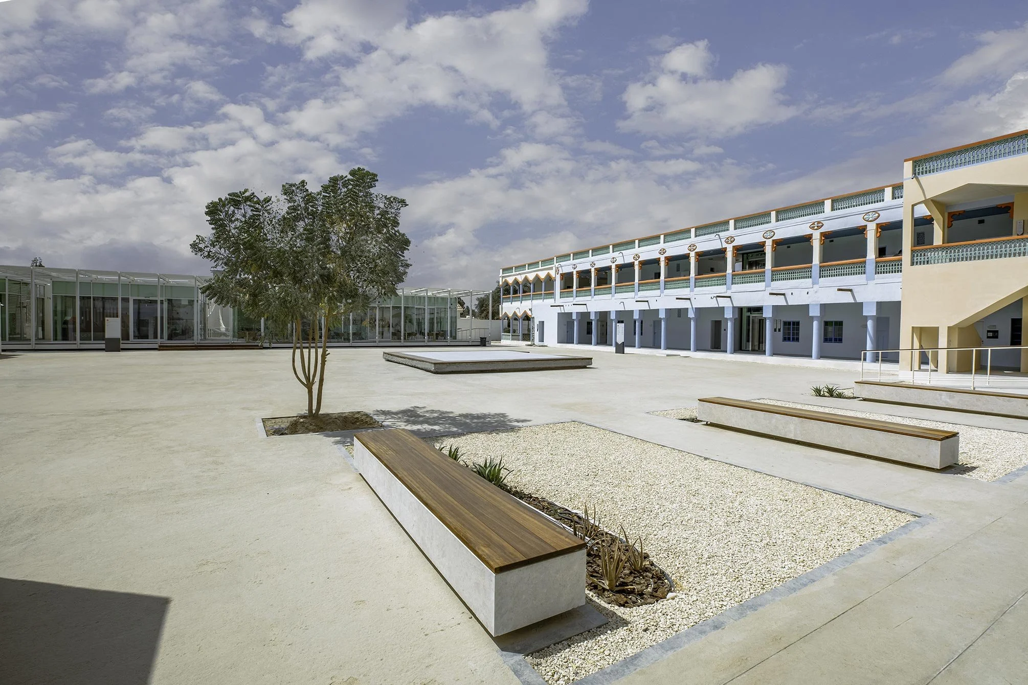 Empty courtyard with benches, a small tree, and modern buildings under a partly cloudy sky.