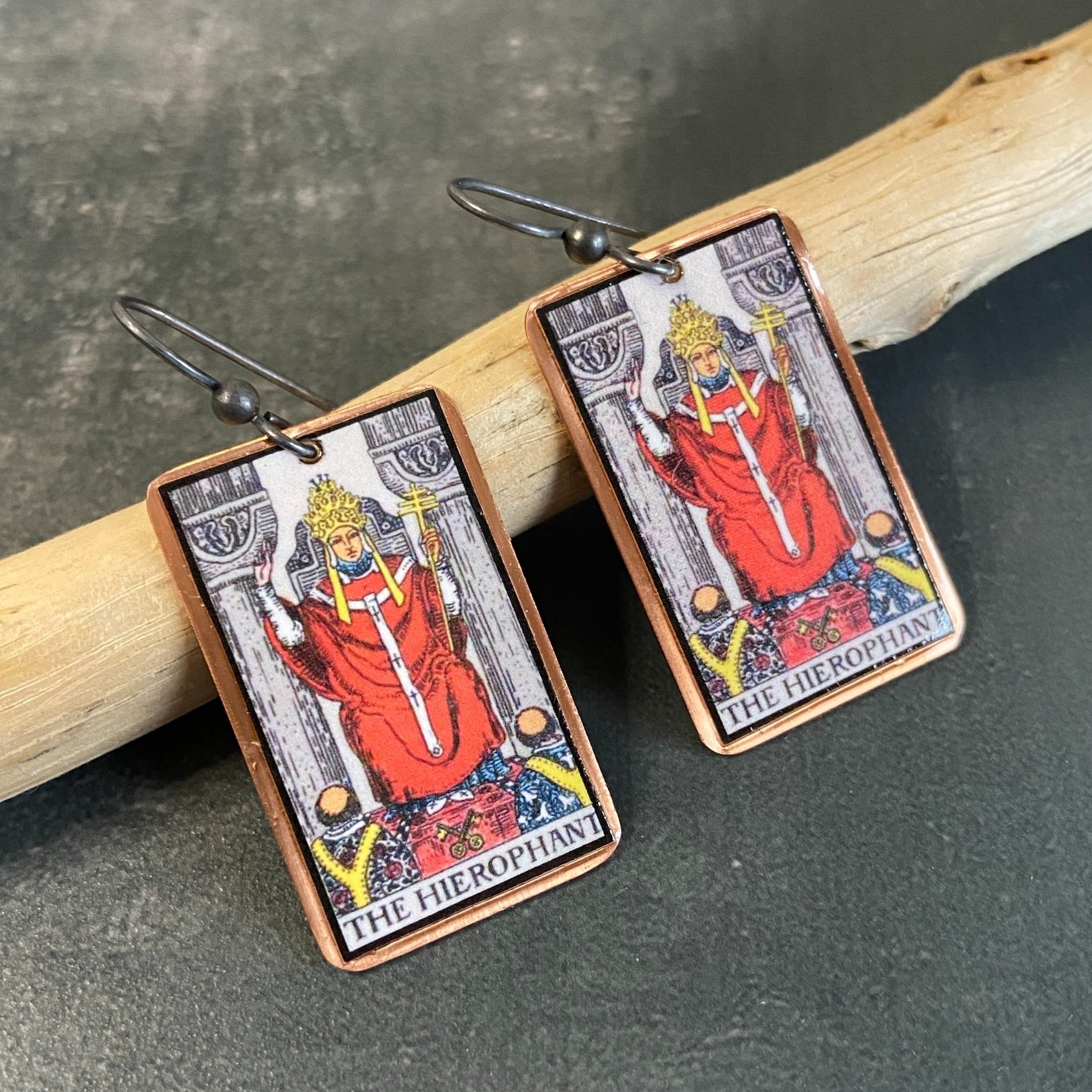 Hierophant Major Arcana tarot earrings, tarot card on copper background with sterling silver ear wires given a dark patina. Earrings learning on drift wood with gray background.