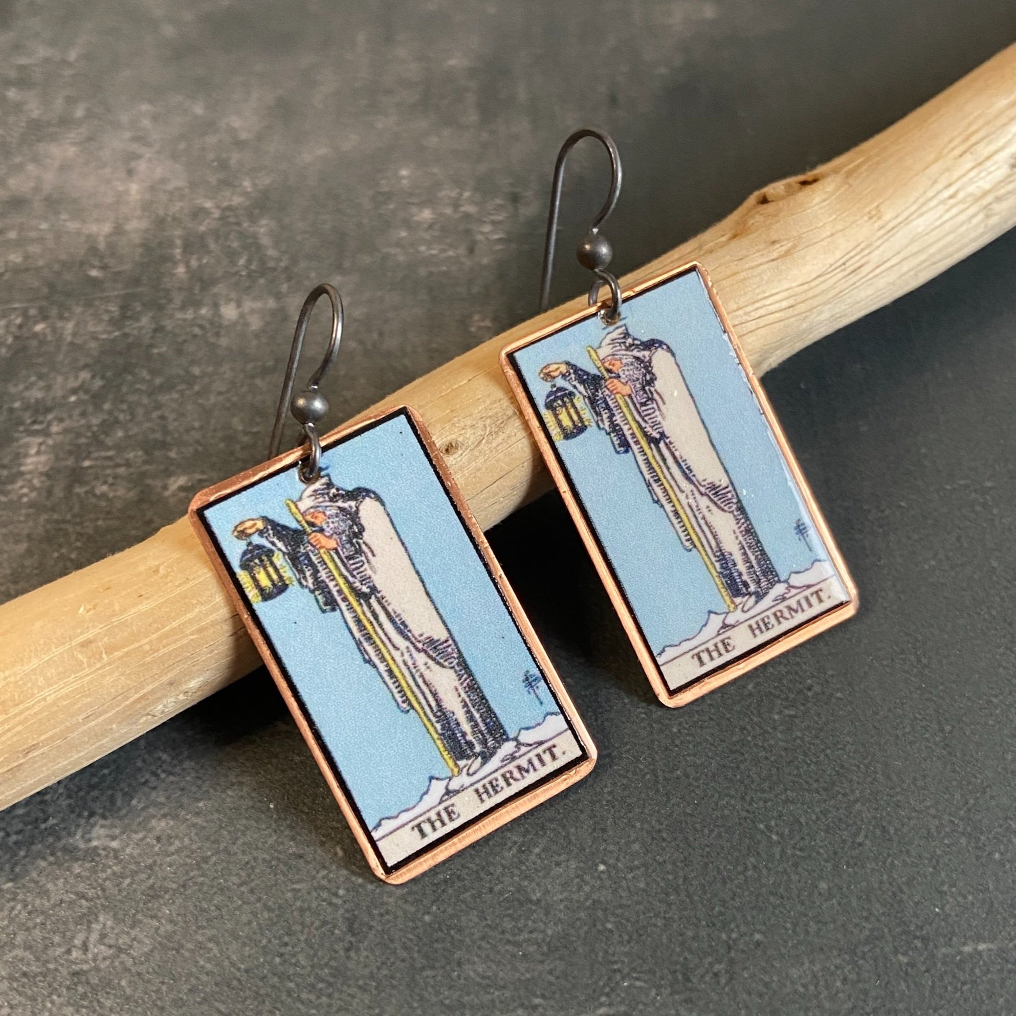 Hermit Major Arcana tarot earrings, tarot card on copper background with sterling silver ear wires given a dark patina. Earrings learning on drift wood with gray background.