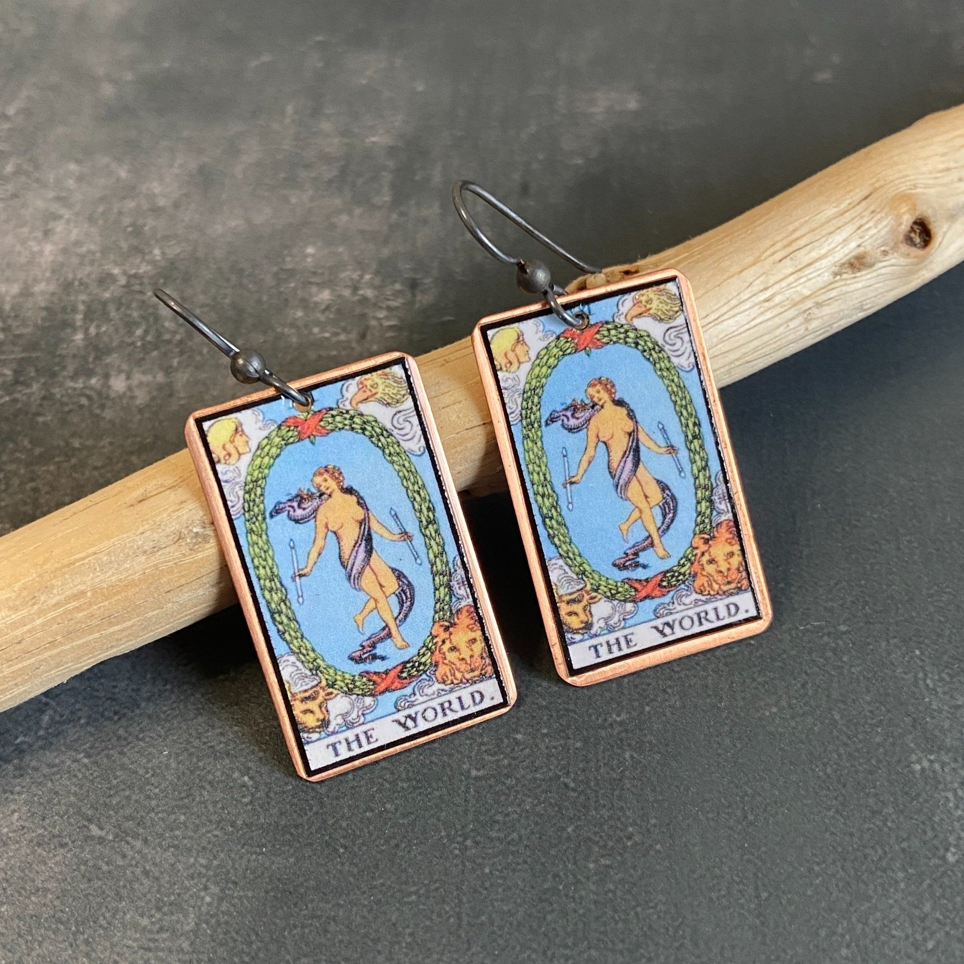 The World Major Arcana tarot earrings, tarot card on copper background with sterling silver ear wires given a dark patina. Earrings learning on drift wood with gray background.