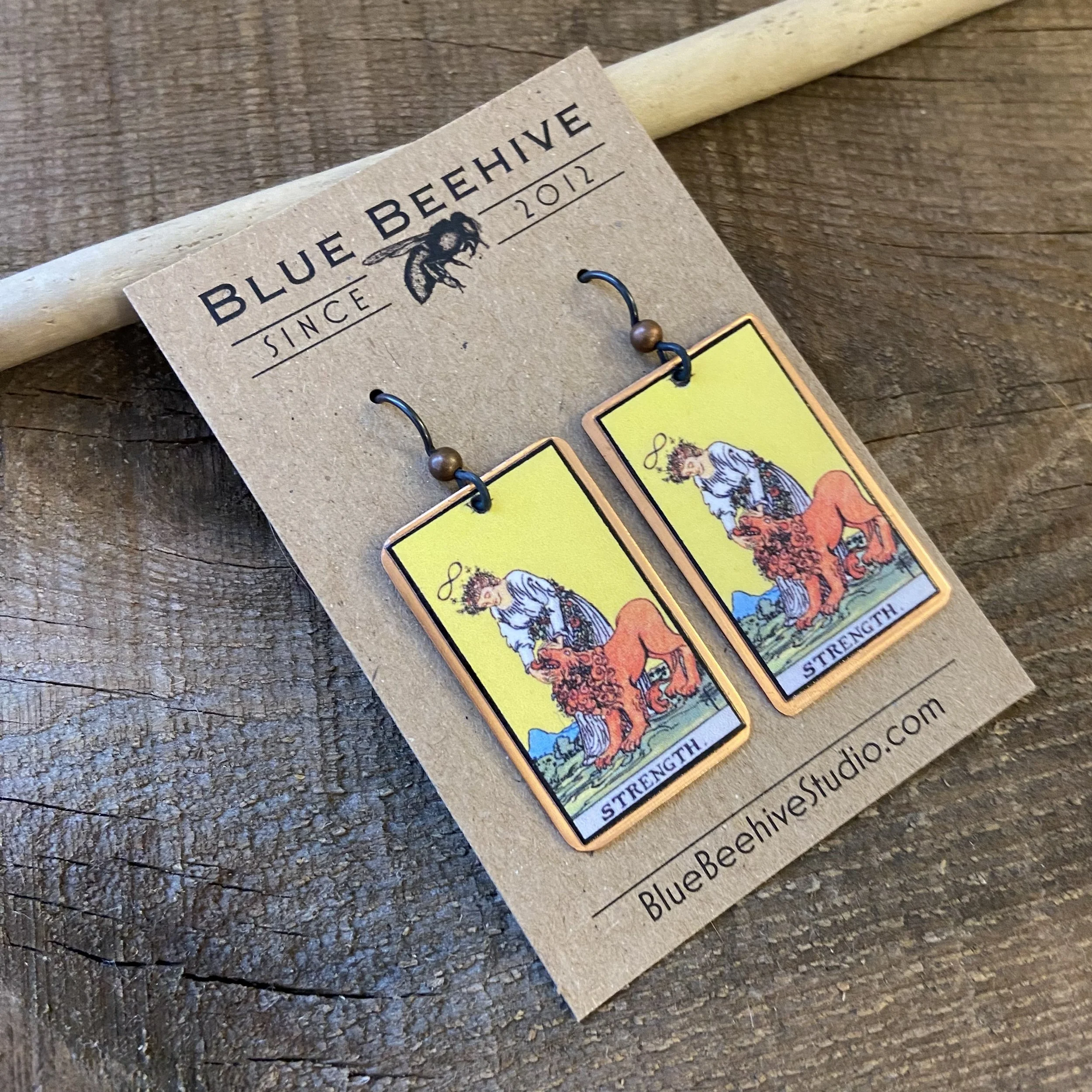 Picture of rectangular earrings of Strength tarot card from the Rider-Waite Deck. Ear wires are black niobium with small copper balls.