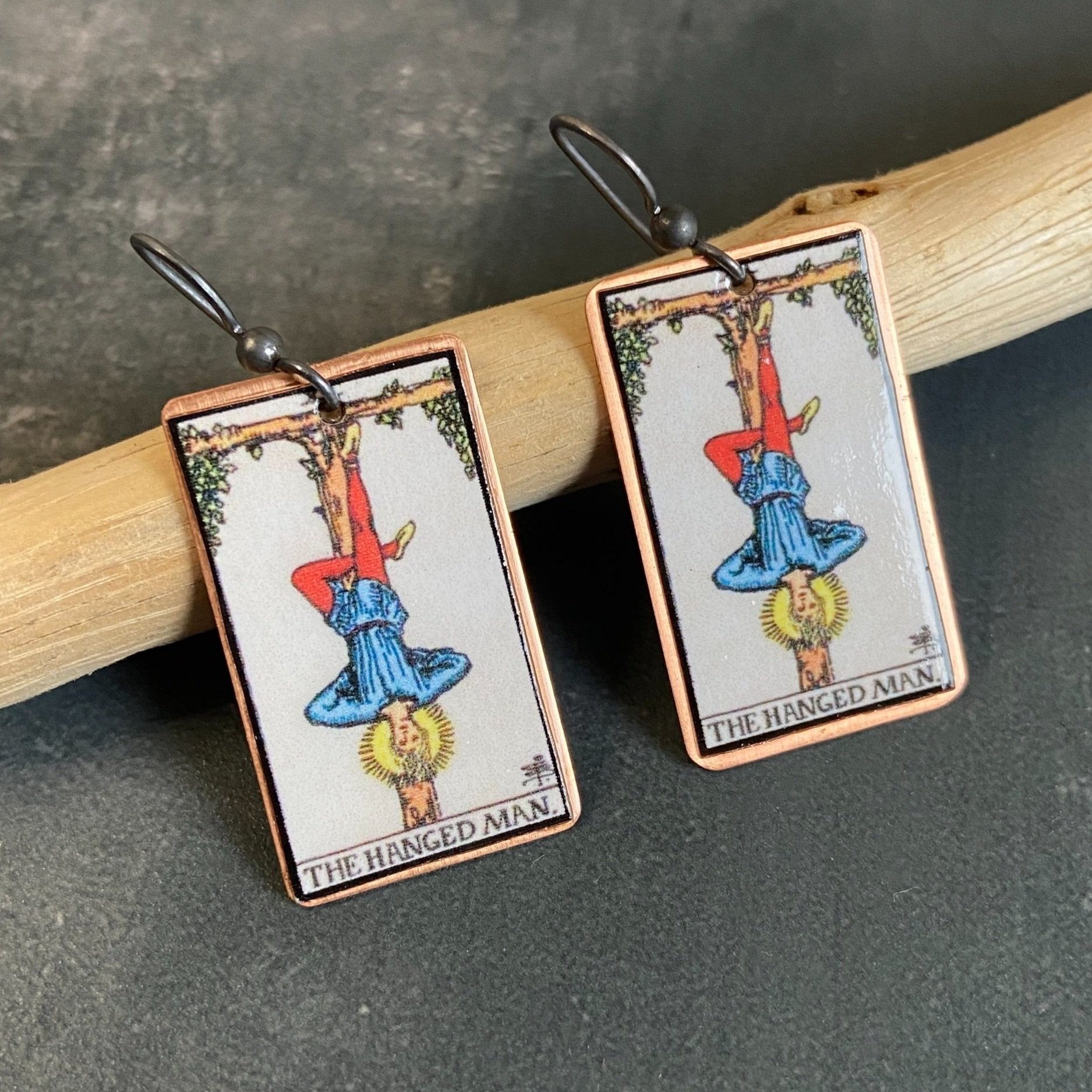 Hanged Man Major Arcana tarot earrings, tarot card on copper background with sterling silver ear wires given a dark patina. Earrings learning on drift wood with gray background.