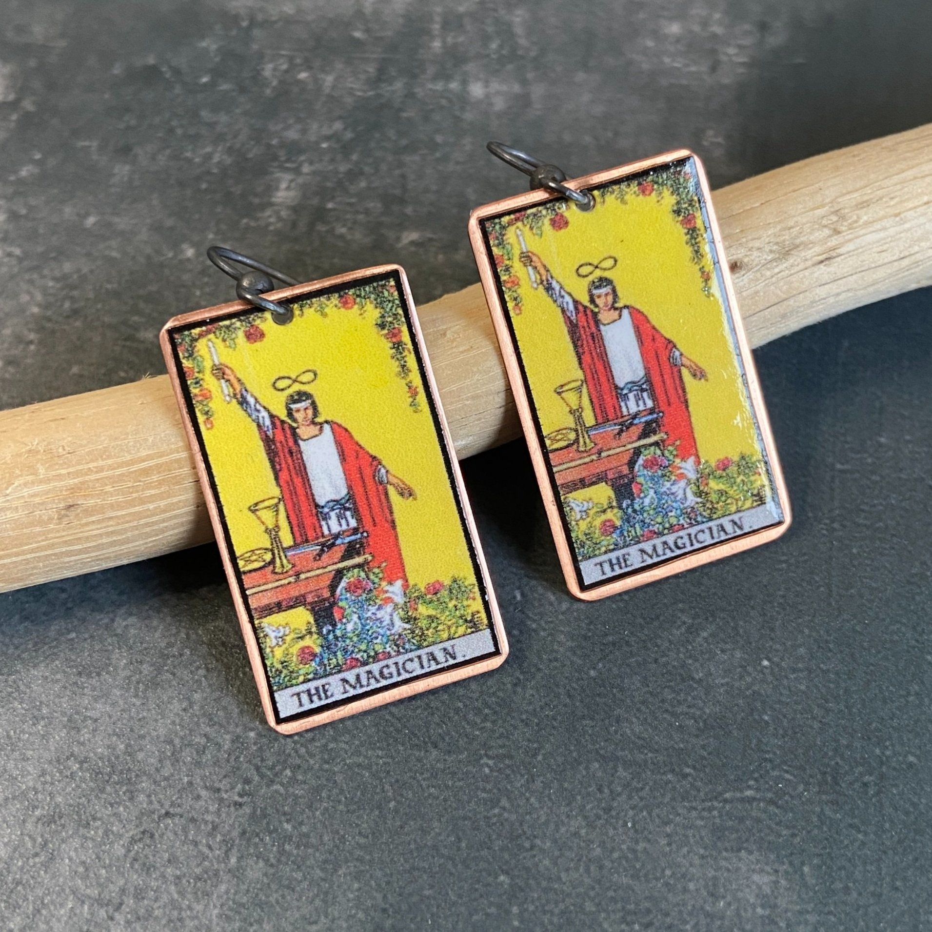 Magician Major Arcana tarot earrings, tarot card on copper background with sterling silver ear wires given a dark patina. Earrings learning on drift wood with gray background.