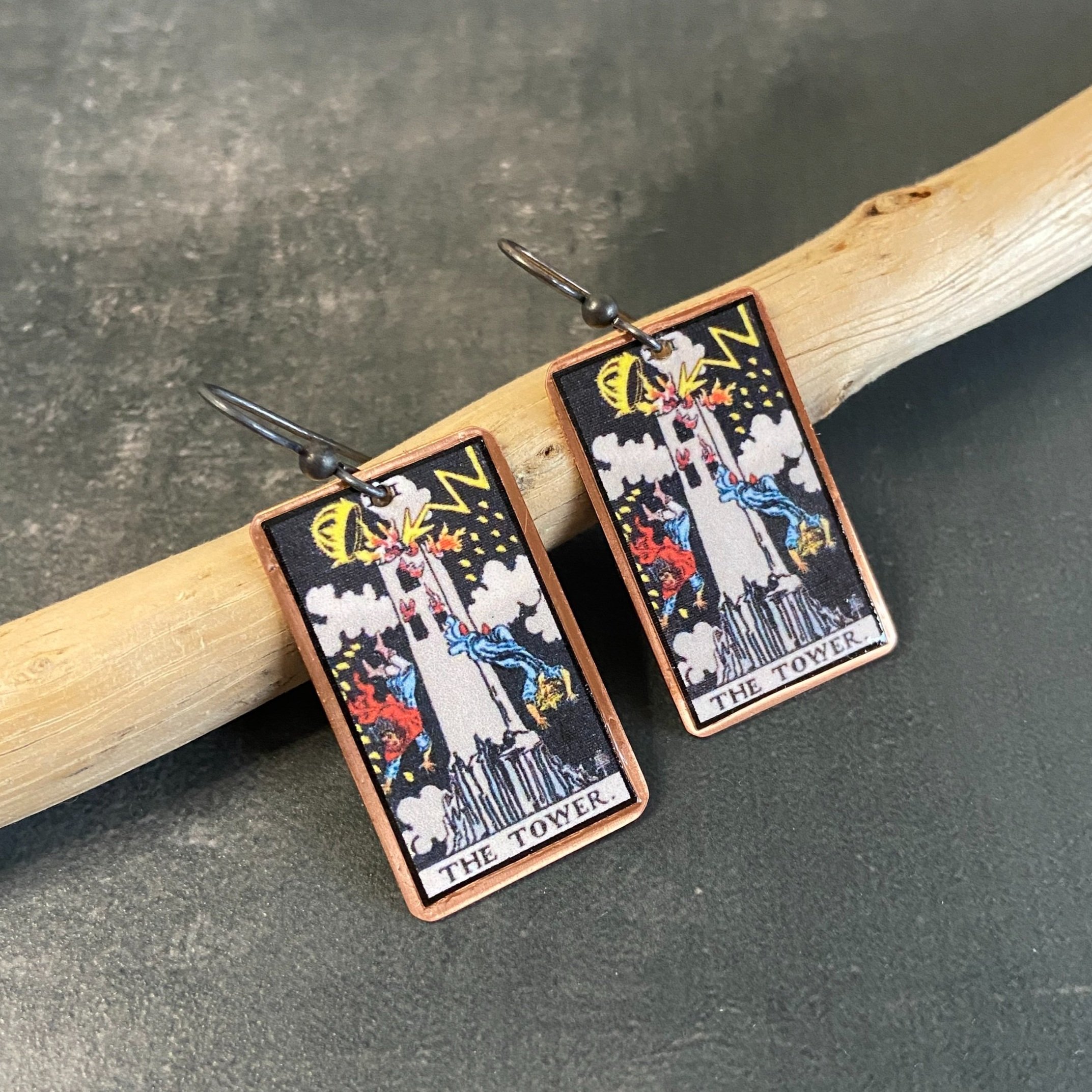 The Tower Major Arcana tarot earrings, tarot card on copper background with sterling silver ear wires given a dark patina. Earrings learning on drift wood with gray background.