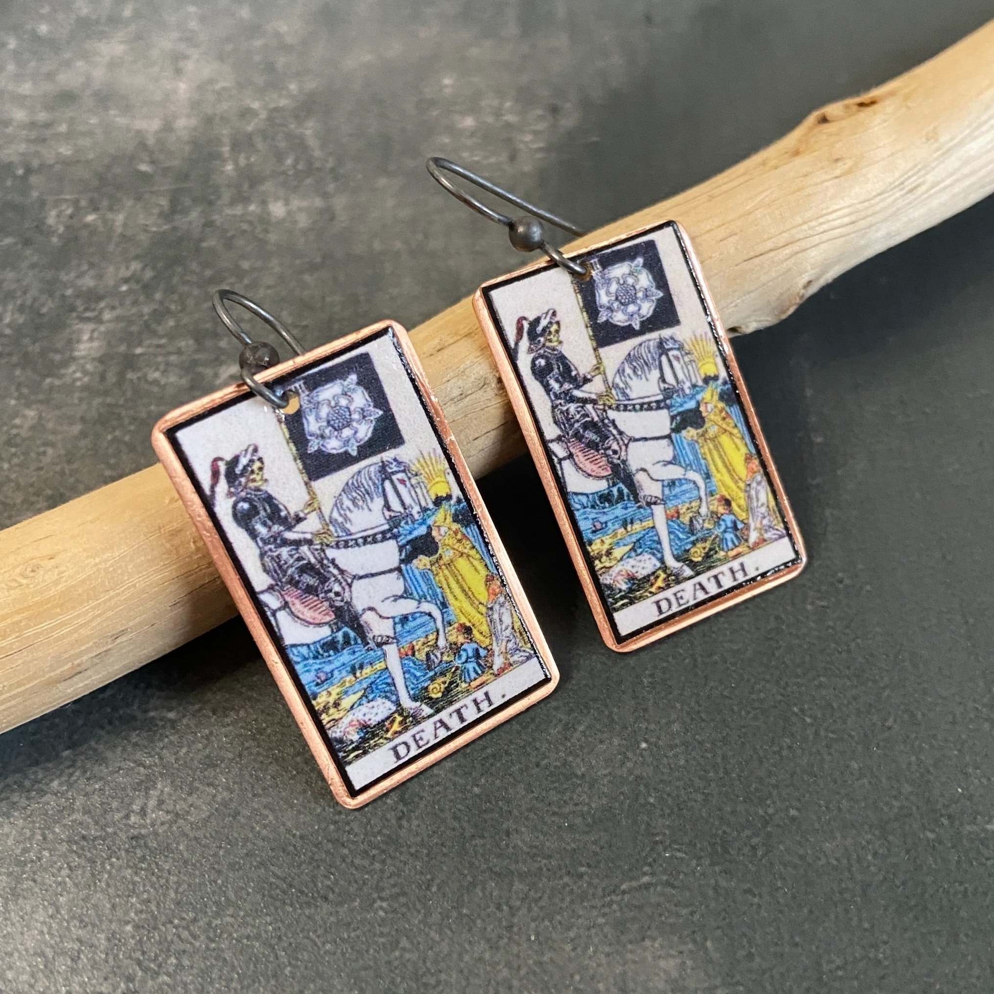 Death Major Arcana tarot earrings, tarot card on copper background with sterling silver ear wires given a dark patina. Earrings learning on drift wood with gray background.