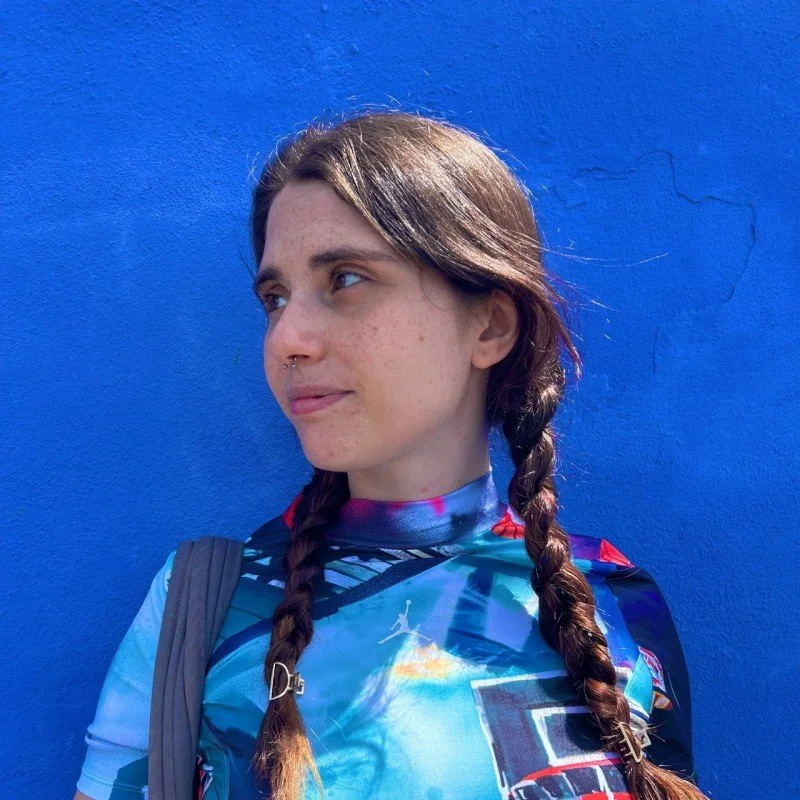 Una ragazza con trecce castane, maglietta sportiva con colori blu e rosso, che porta uno zaino e si trova davanti a una parete blu.