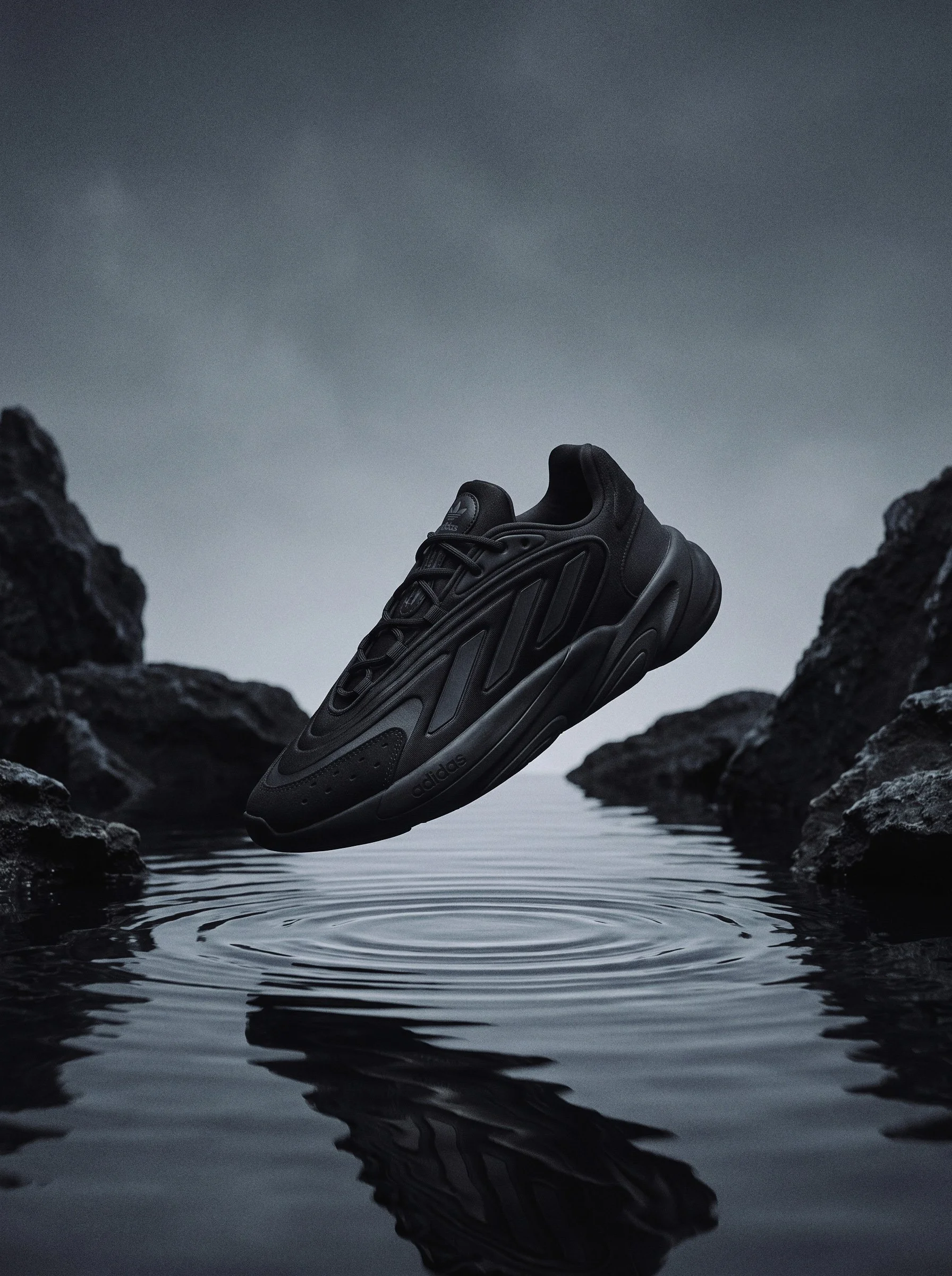A black Adidas sneaker floating above a body of water surrounded by rocks with a cloudy sky in the background.