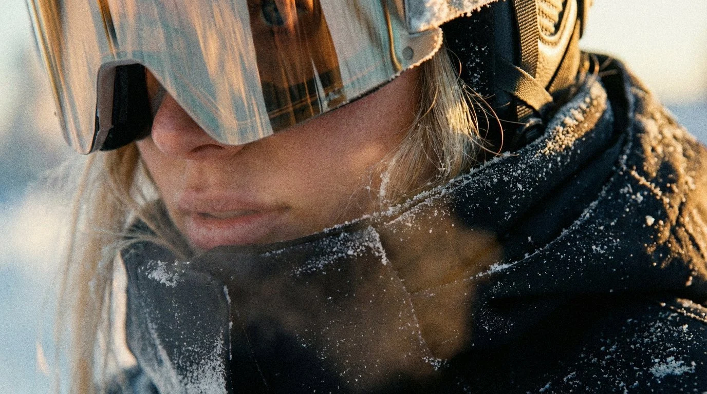 Close-up portrait of a young woman with natural freckles, long dark hair, and a silver nose ring, gazing intensely toward the camera. Soft, cinematic lighting with a blurred foreground featuring a sleek metallic surface accented by orange highlights.