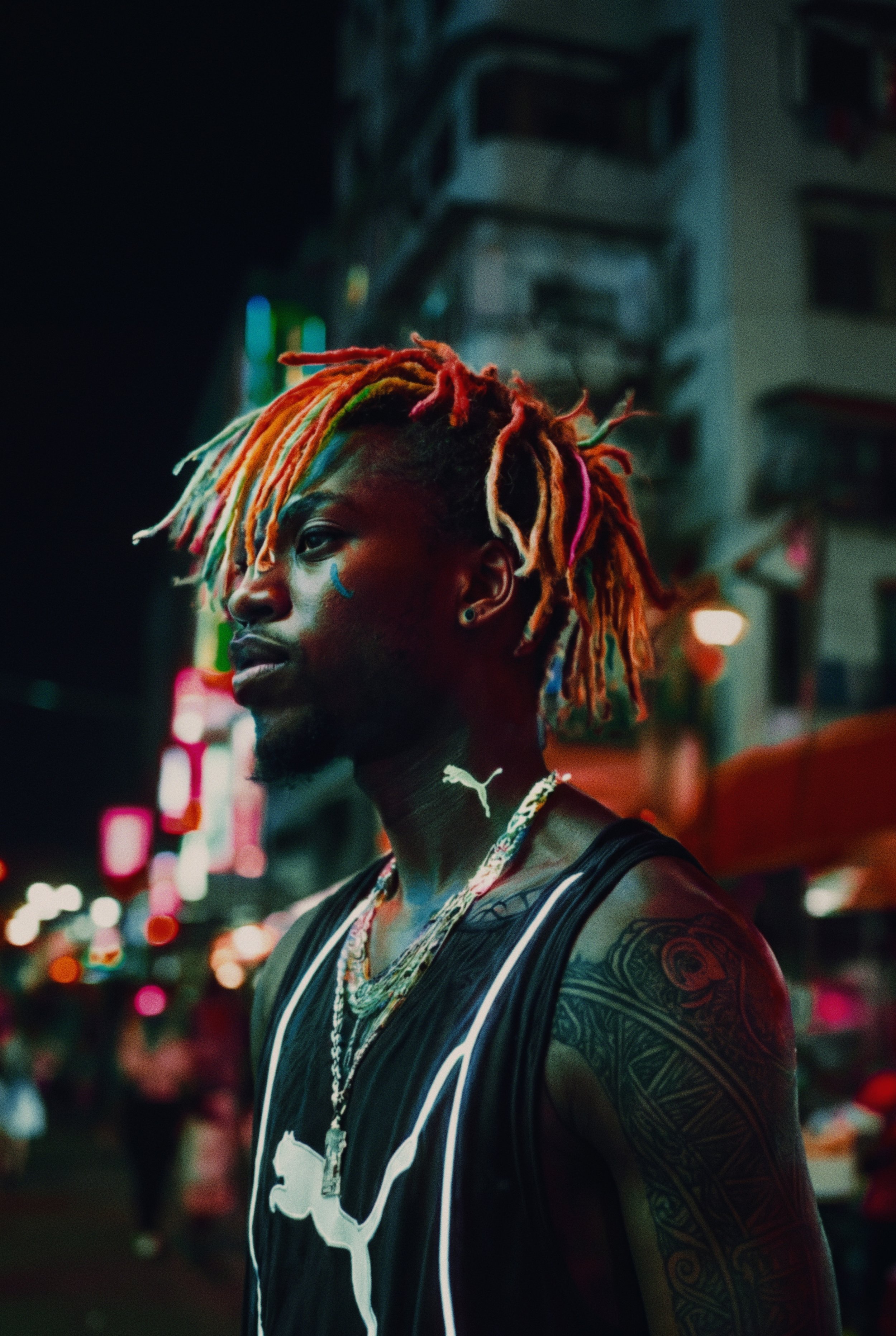 A person with multi-colored dreadlocks and tattoos on their arm, standing on a city street at night with blurred lights in the background.