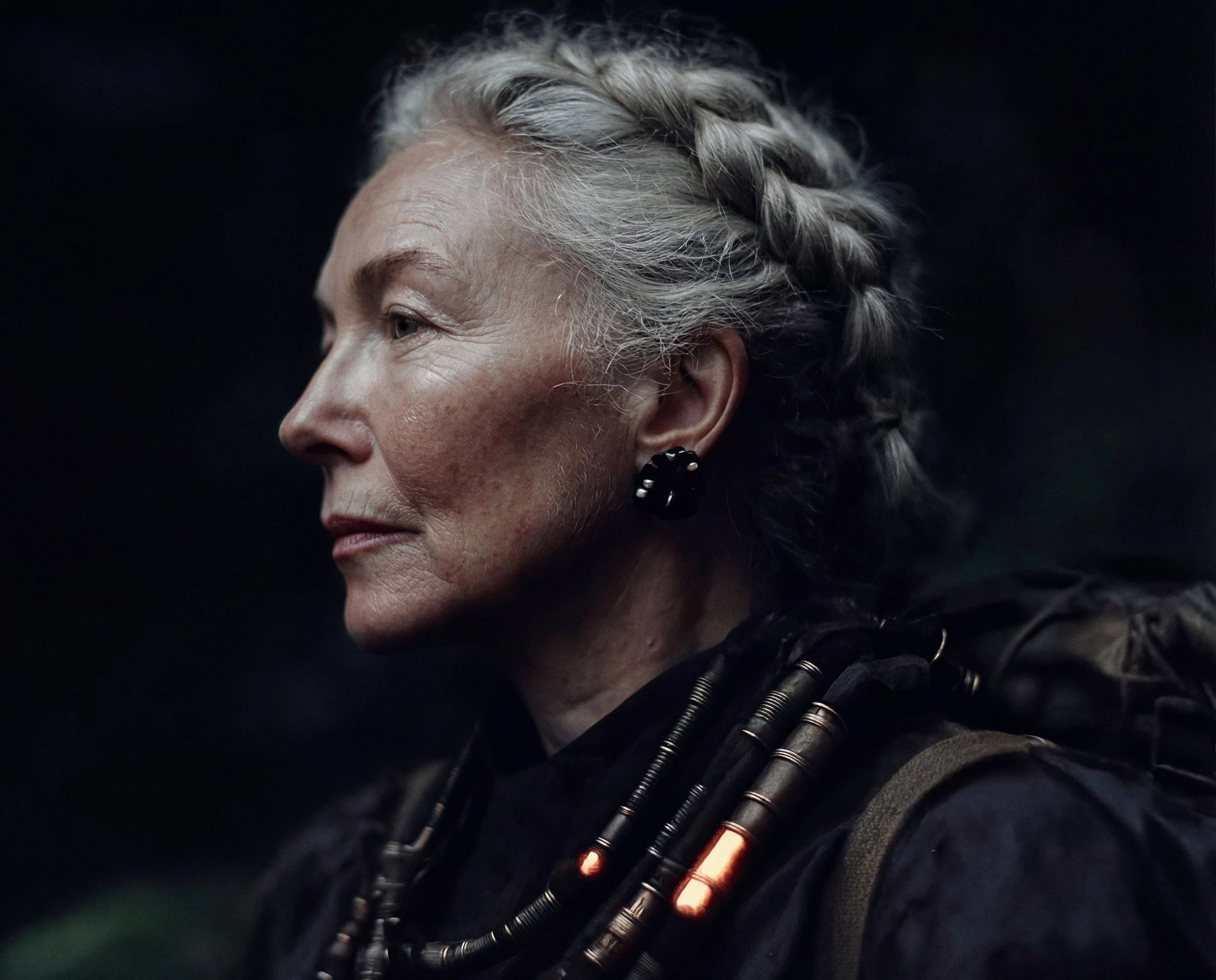 Close-up portrait of a young woman with natural freckles, long dark hair, and a silver nose ring, gazing intensely toward the camera. Soft, cinematic lighting with a blurred foreground featuring a sleek metallic surface accented by orange highlights.