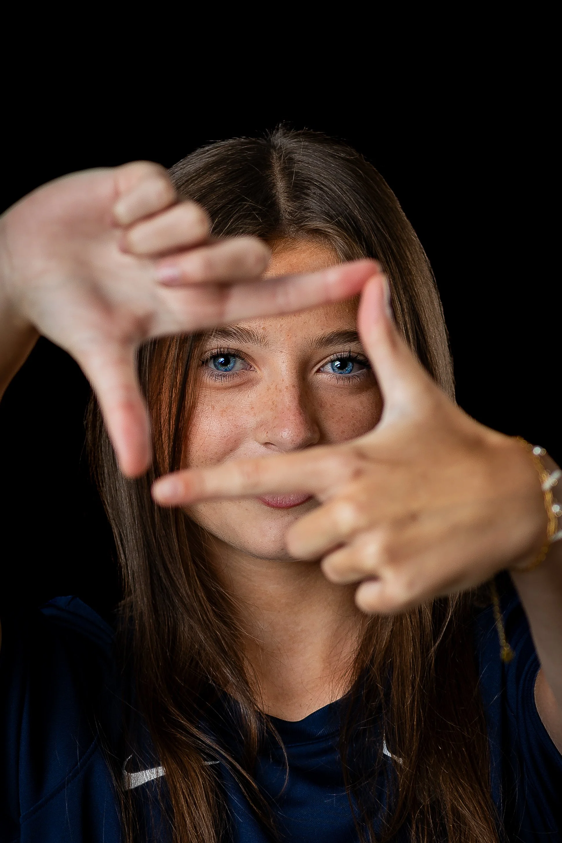 A business headshot portrait of a girl with blue eyes and freckles looking through her own frame made from her first finger and thumb of both hands back at the camera
