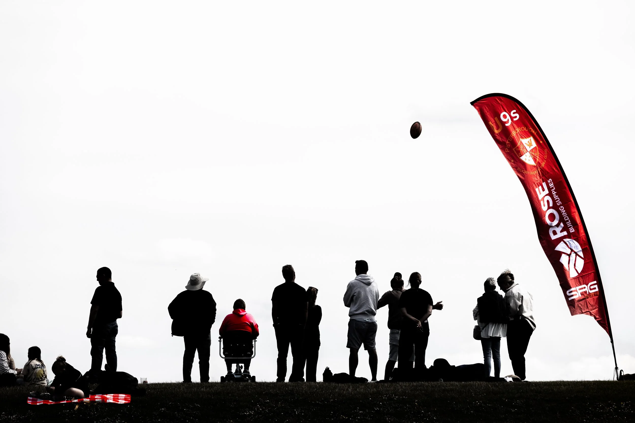 The sponsors banner flies in the wind dominating the skyline next to a rugby pitch as the spectators look on at the match silhouetted by the sky