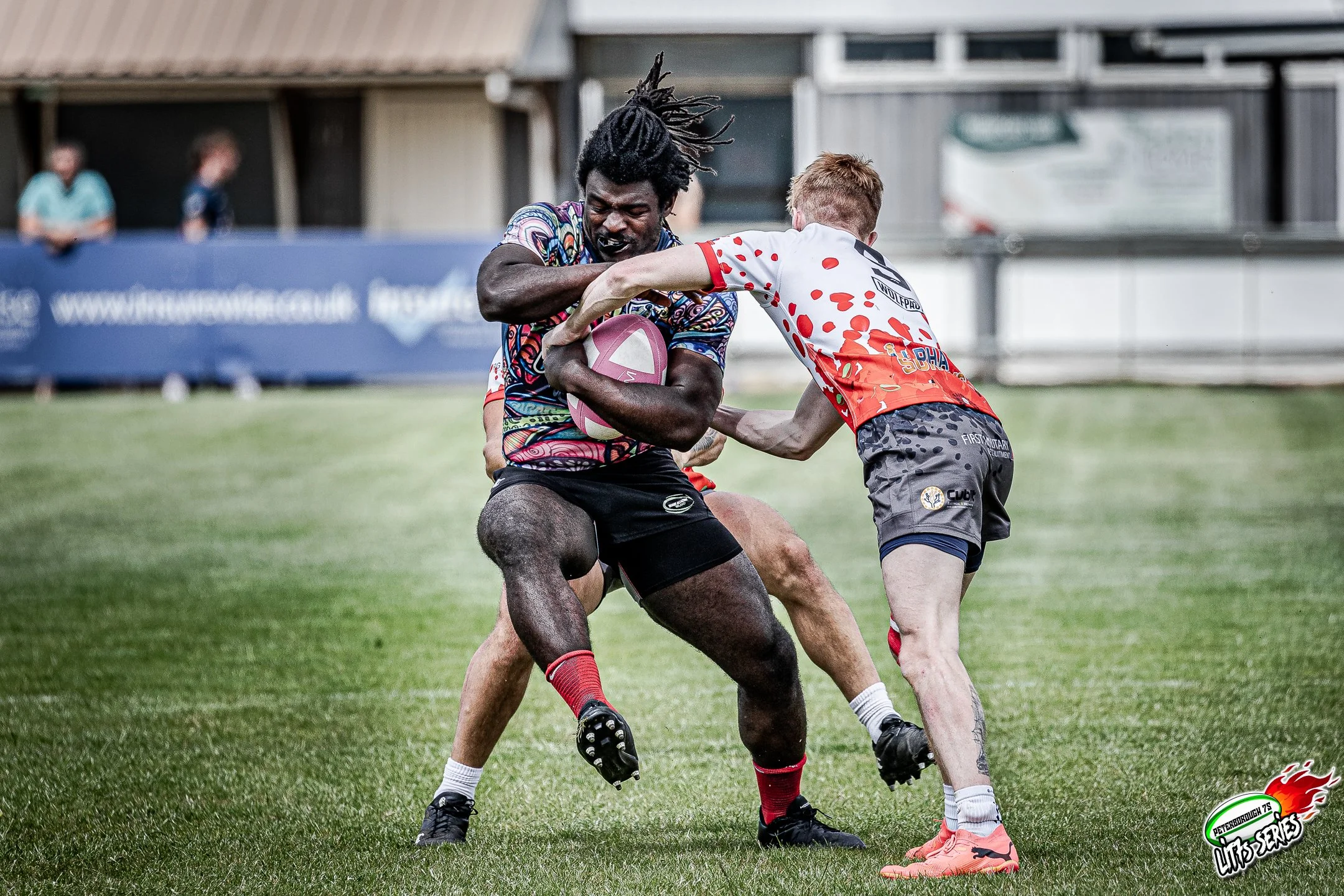 Two rugby players competing for the ball, one holding the ball tightly, while the other tries to grab it, on a grassy field with spectators and banners in the background.