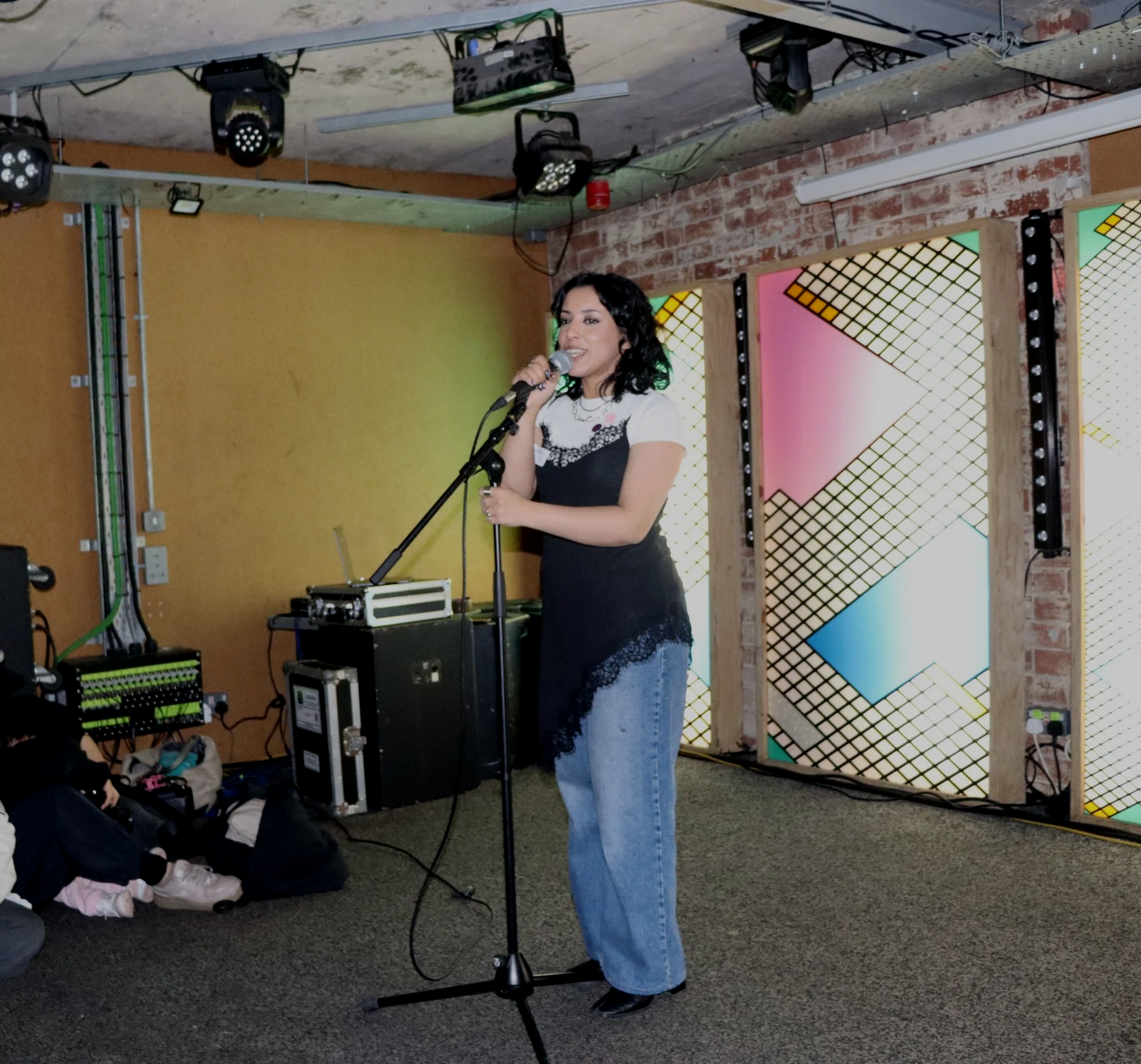 A woman standing in front of an audience with a microphone, wearing a white and black top and loose jeans, with colourful geometric art on the wall.