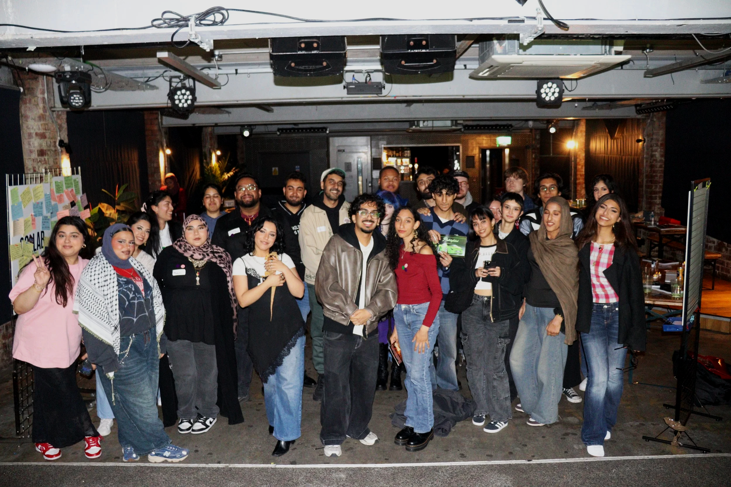 A large group of diverse people posing for a photo in an indoor venue, with brick walls, stage lights, and some decor in the background.
