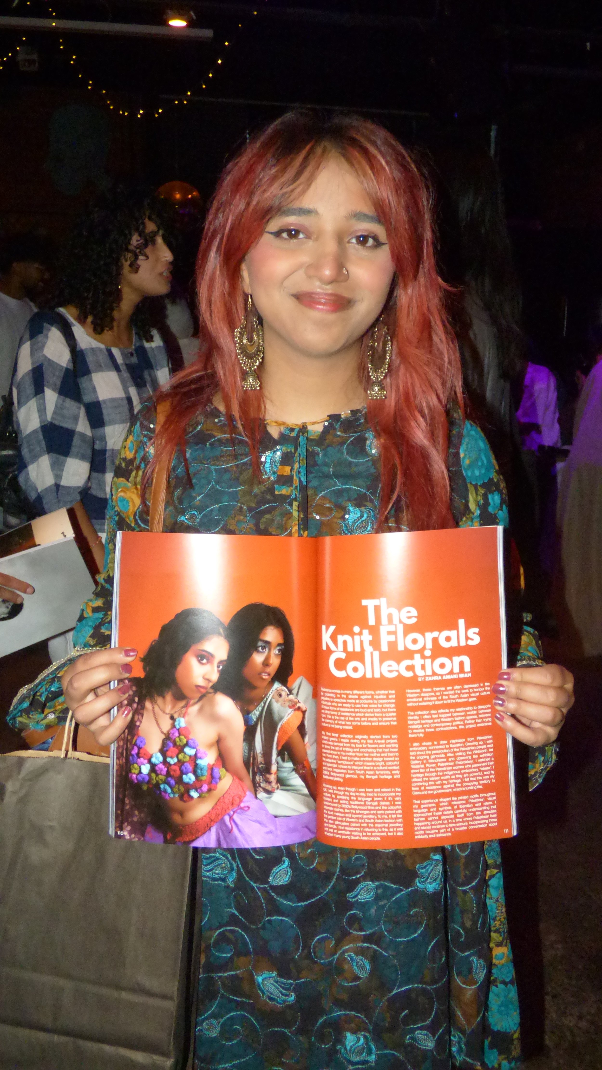 A woman with red hair and earrings holding an open magazine titled 'The Knit Florals Collection' at an indoor event with dark lighting and people in the background.