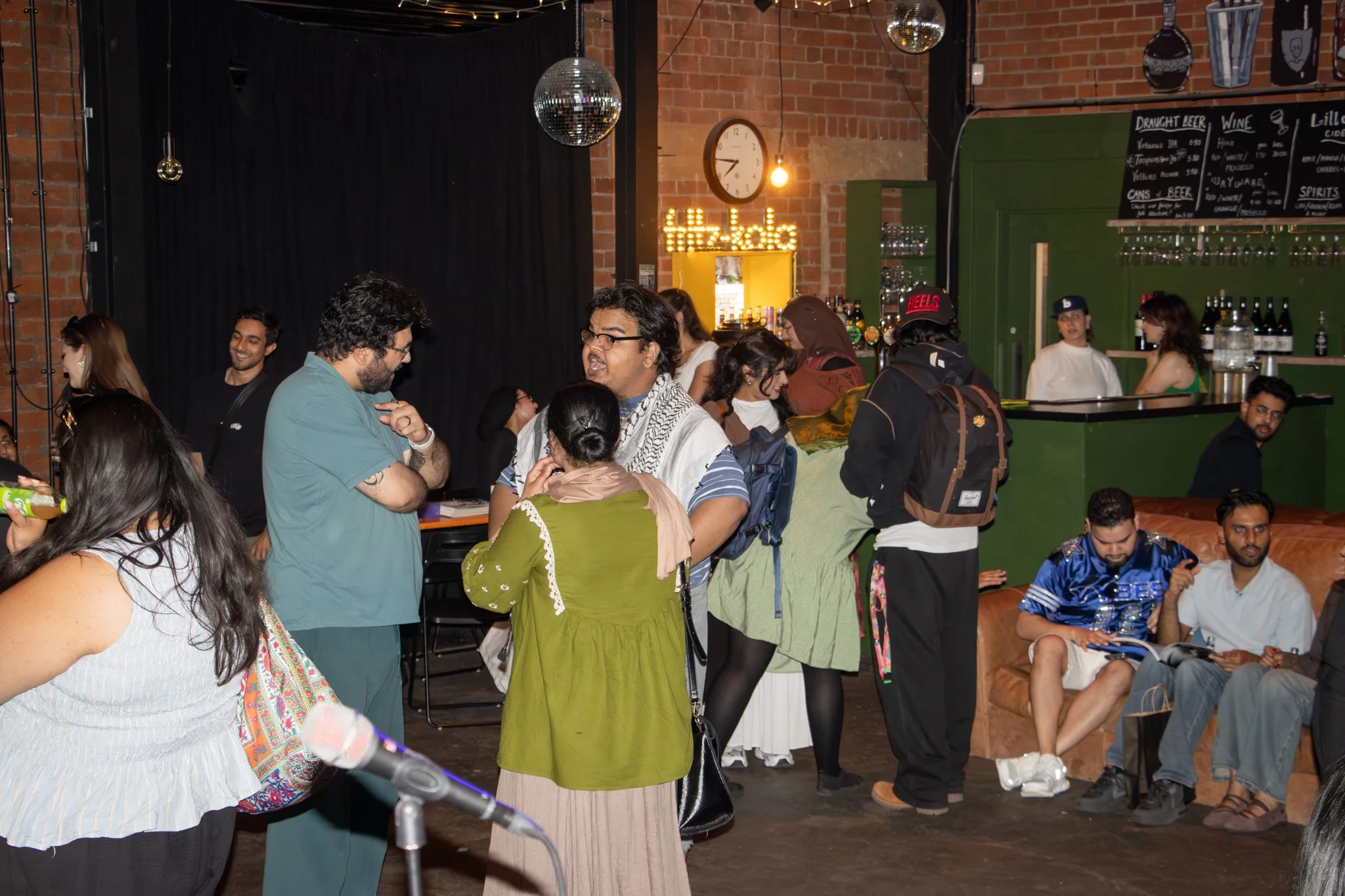 A lively indoor gathering of diverse adults socializing, some standing and chatting, others sitting on couches, with a bar area in the background and disco balls hanging from the ceiling, illuminated by warm lighting.