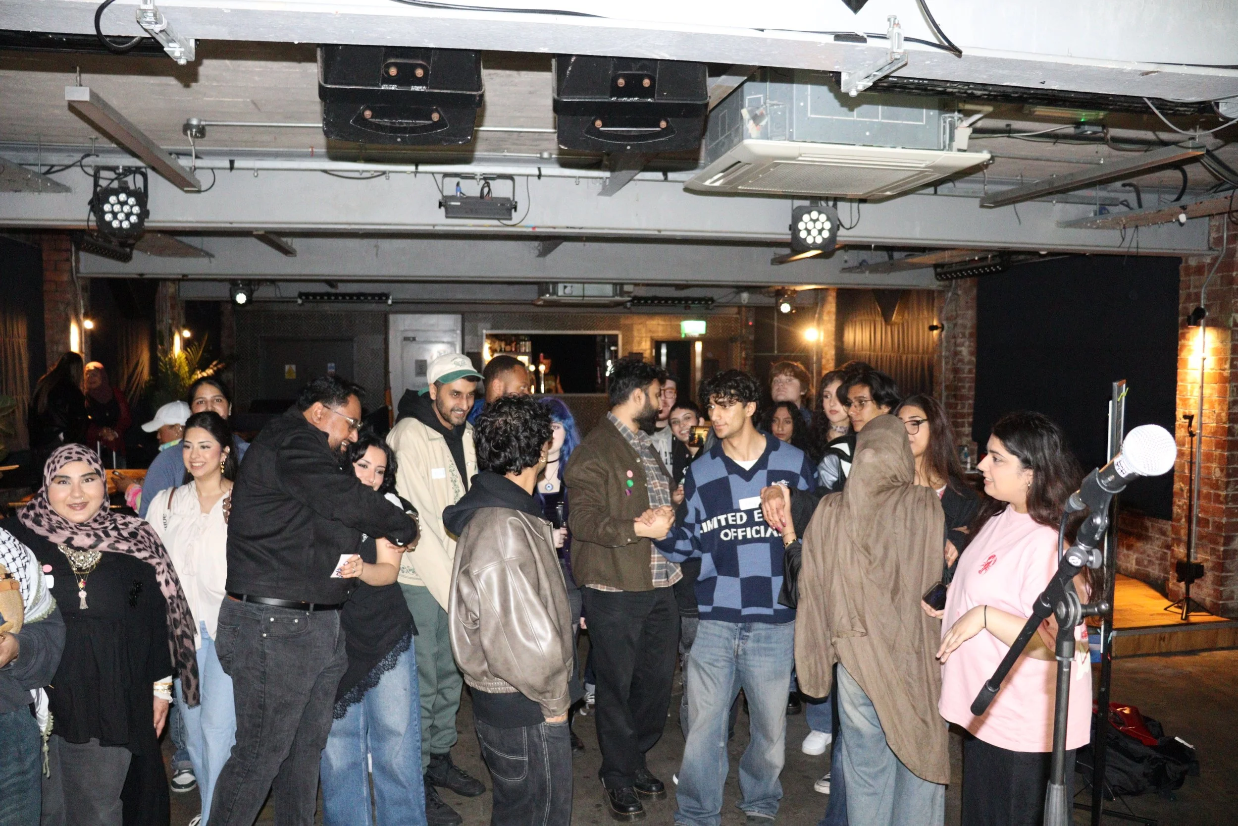 People gathered in a crowded indoor space, engaging in conversations. The room has exposed brick walls, industrial lighting, and hanging speakers and equipment on the ceiling. Some individuals are smiling and interacting with each other.