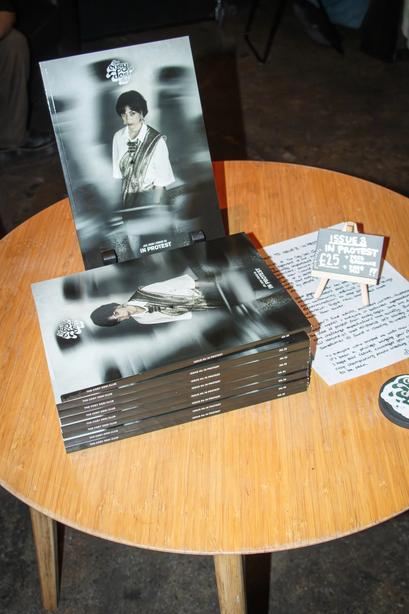 A stack of magazines on a wooden table. The magazine's title is 'The Cosy Desi Club' and the issue is  'In Protest'. 