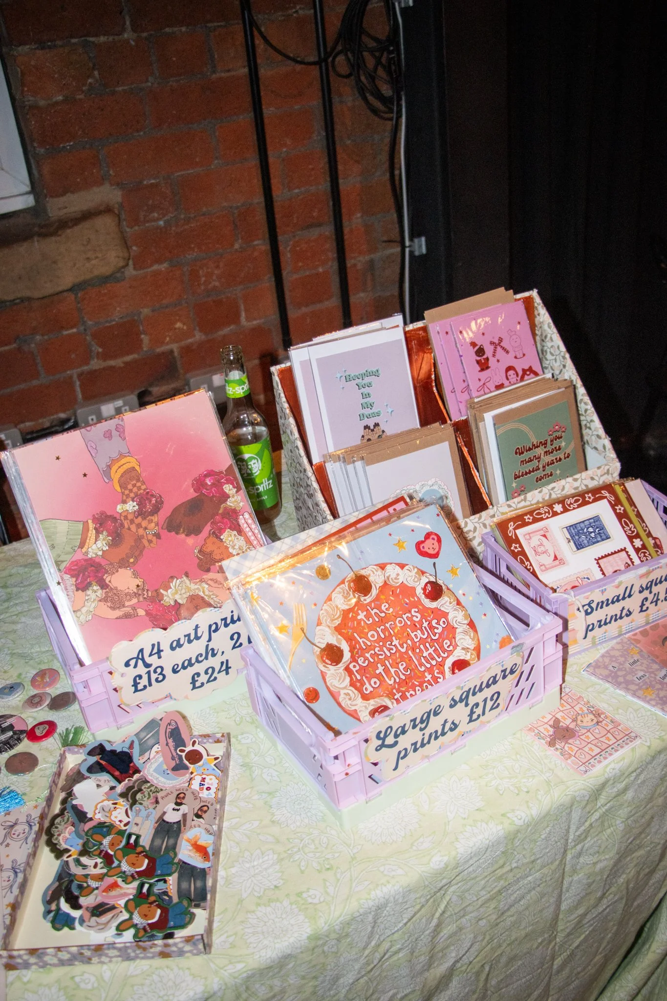 A display of colorful greeting cards and stationery on a table with a green and white patterned tablecloth, against a brick wall background.