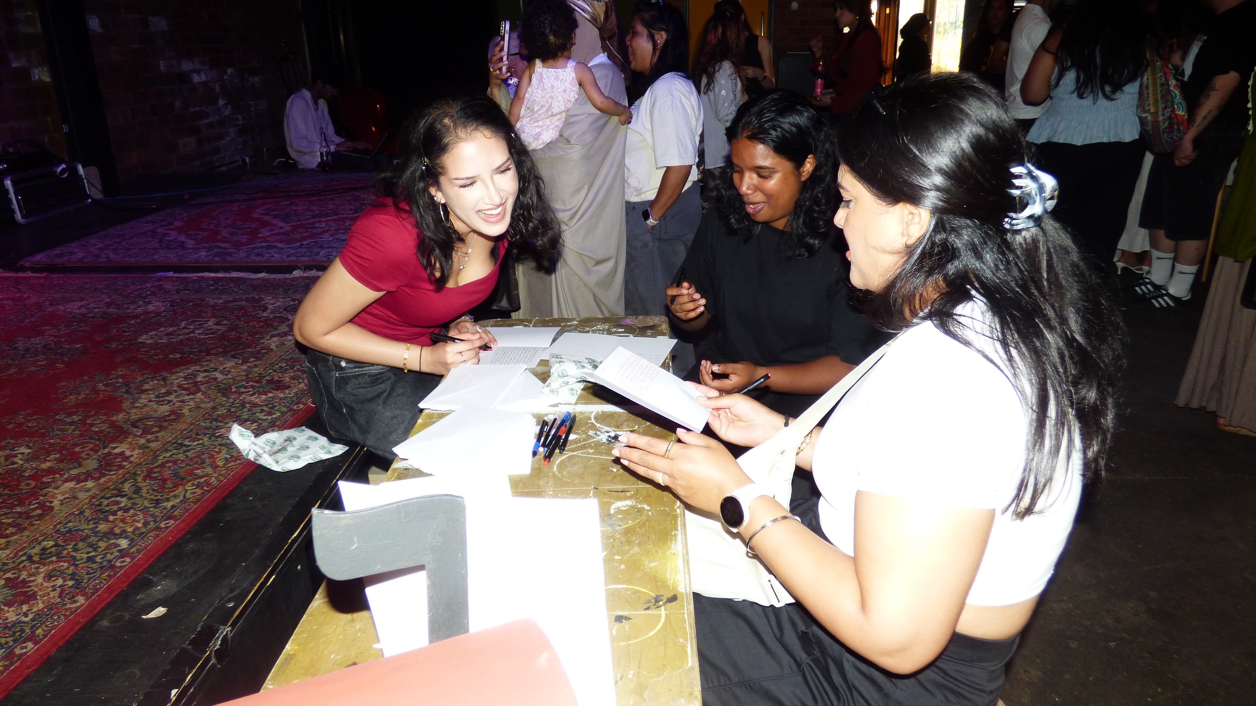 Three people sitting at a table with papers and pens, engaging in conversation at an indoor event with a brick wall and people in the background.