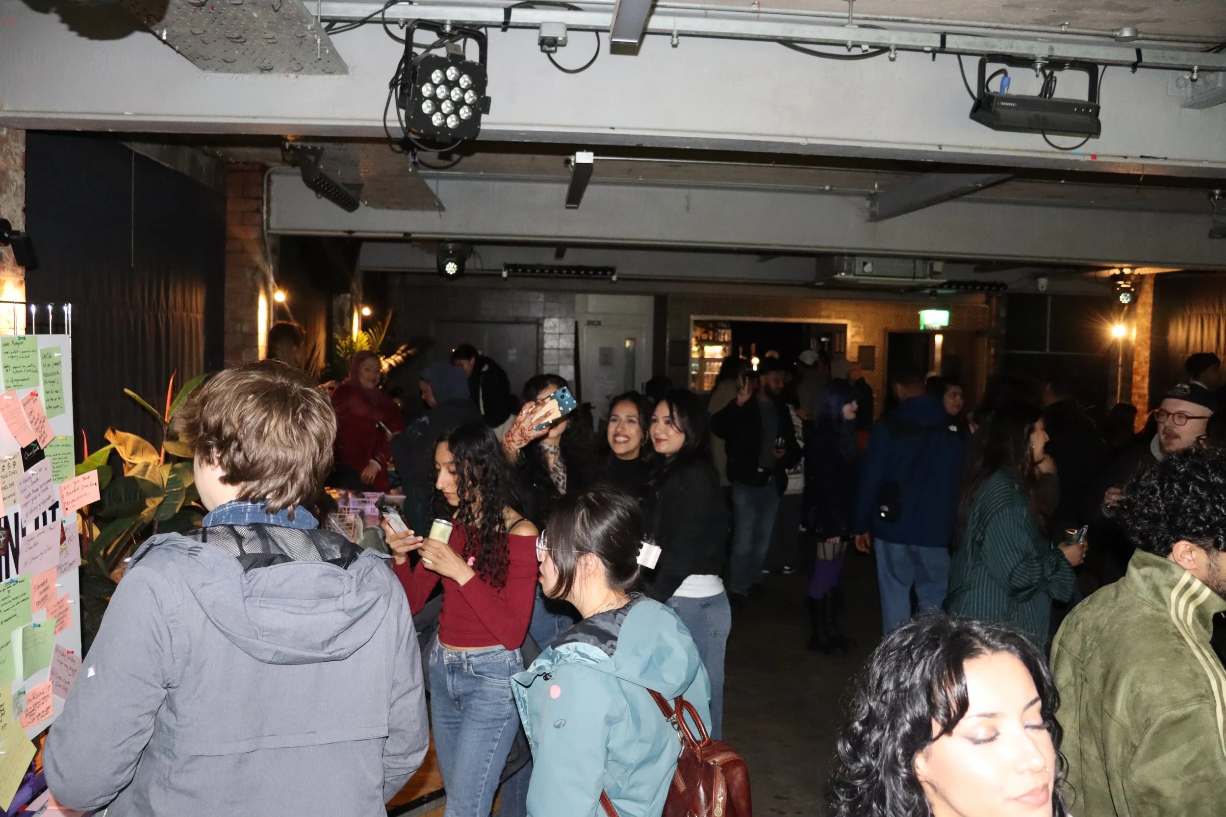 Crowd of people socialising in a dimly lit indoor space with exposed ceiling beams, lighting fixtures, and a wall with notes or decorations.
