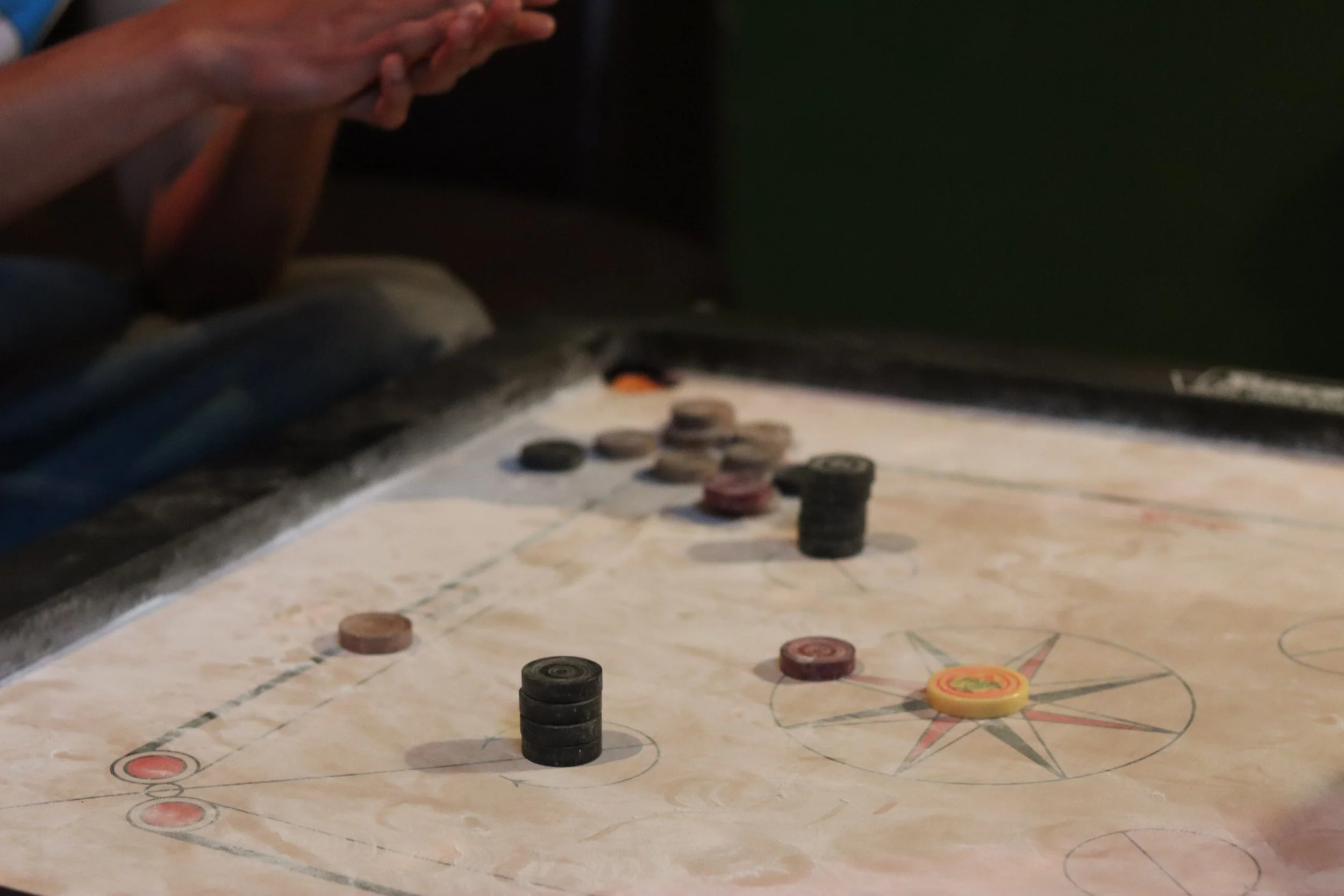 Close-up of a wooden tabletop with scattered black and red game pieces from a board game, with part of a hand visible on the left side and a dark green wall in the background.