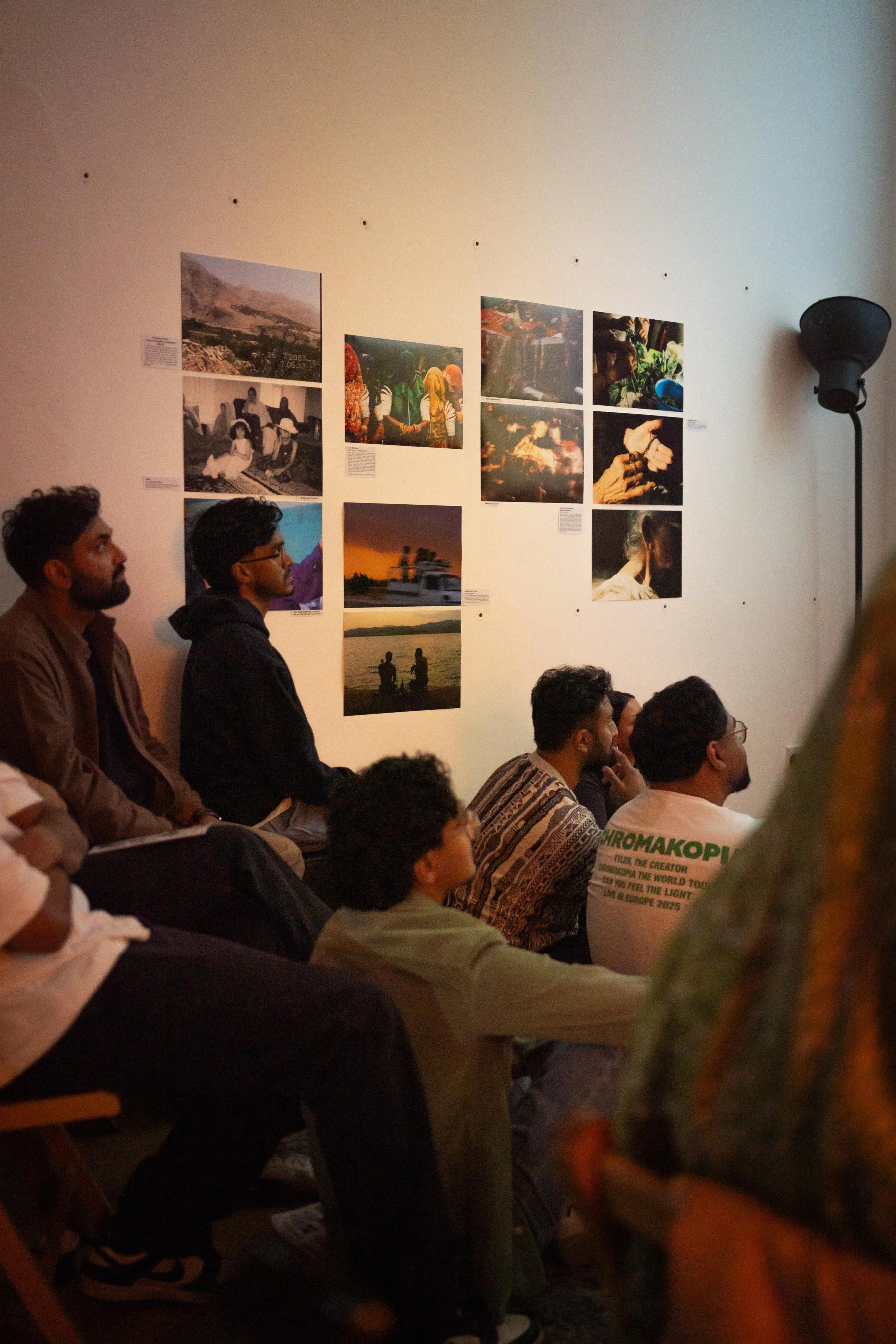 An indoor art exhibition with people seated and looking towards the artwork on the wall, which displays a collection of photographs showing landscapes, people, and scenes of daily life.
