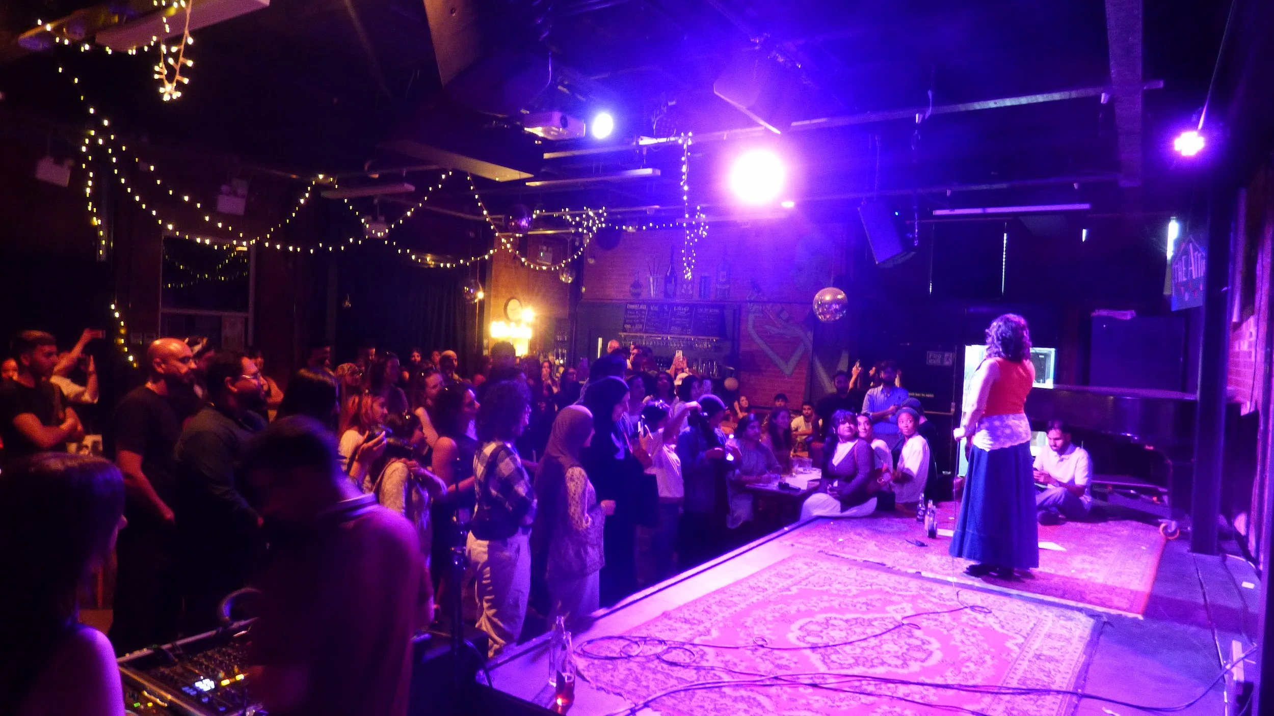 A crowded indoor venue with people sitting and standing, facing a stage where a woman is speaking. The room is decorated with string lights.