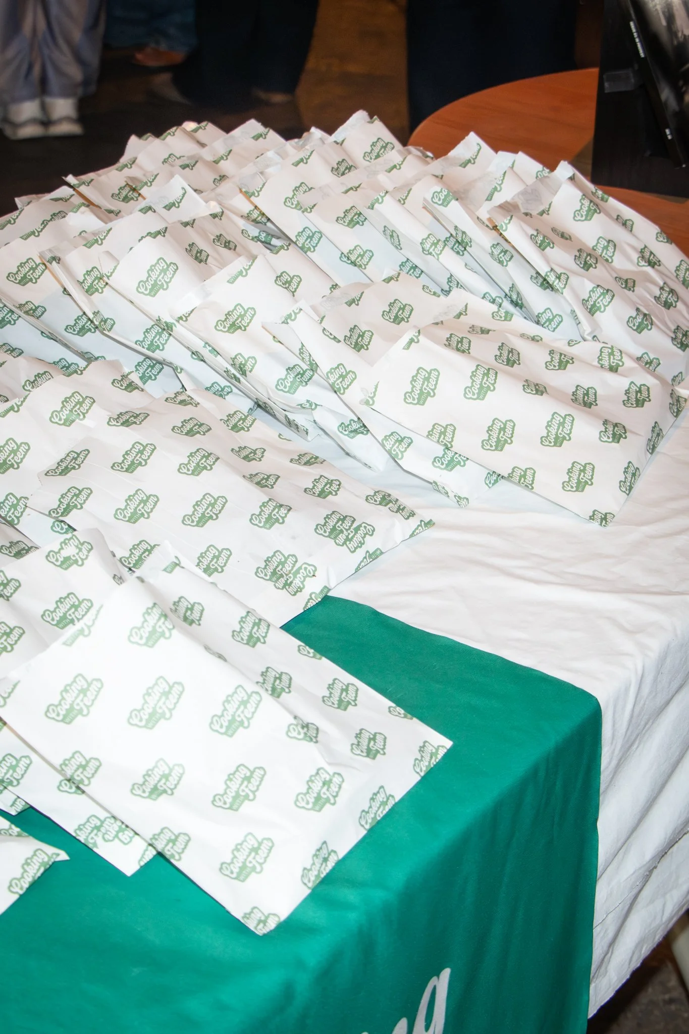 A table covered with white paper bags printed with green text that reads 'Cooking with Feem' and a green tablecloth underneath.