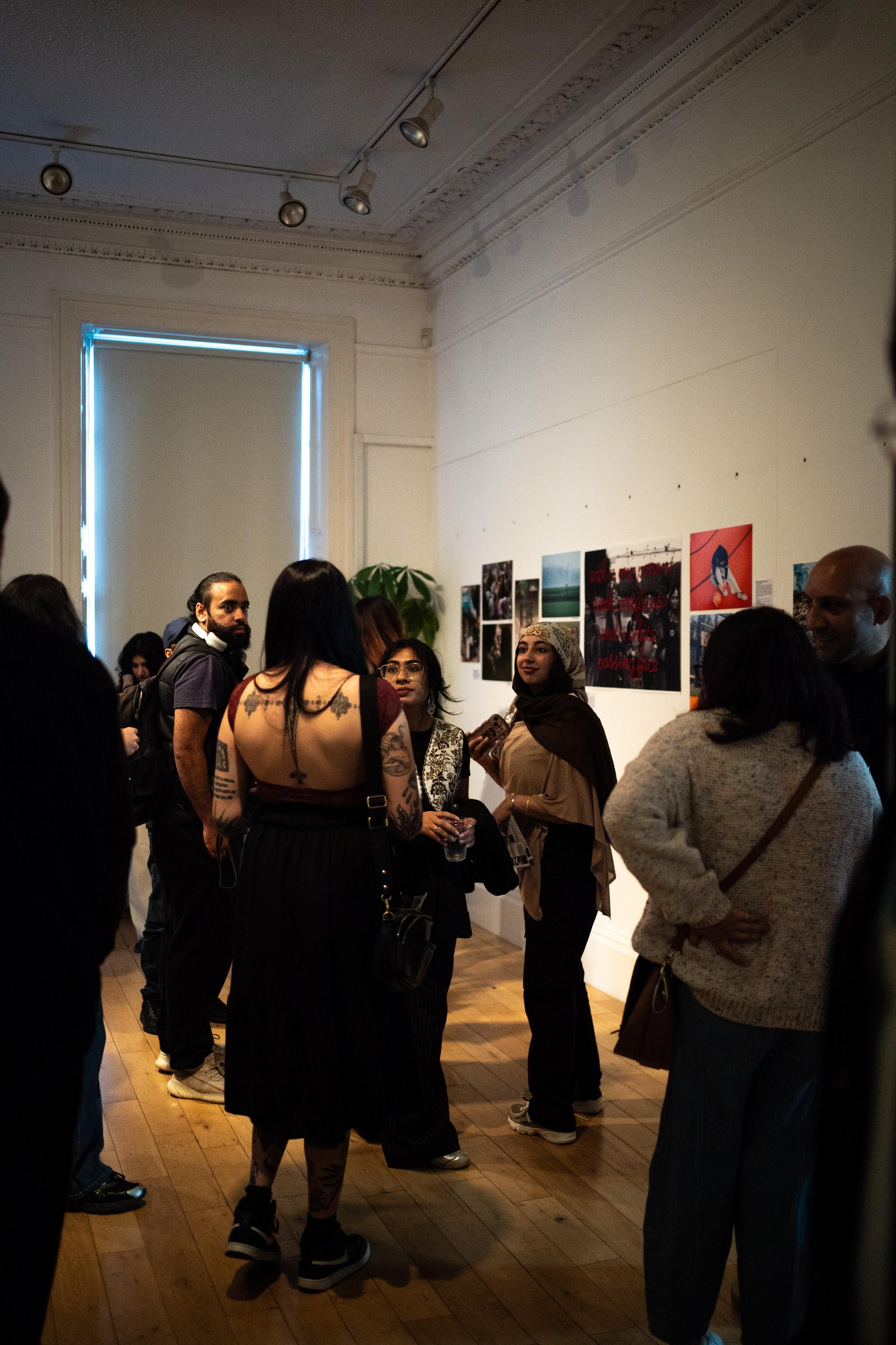A group of people gathered in an art gallery, engaging in conversations. The gallery has photographic artworks displayed on the white walls, and light fixtures on the ceiling.