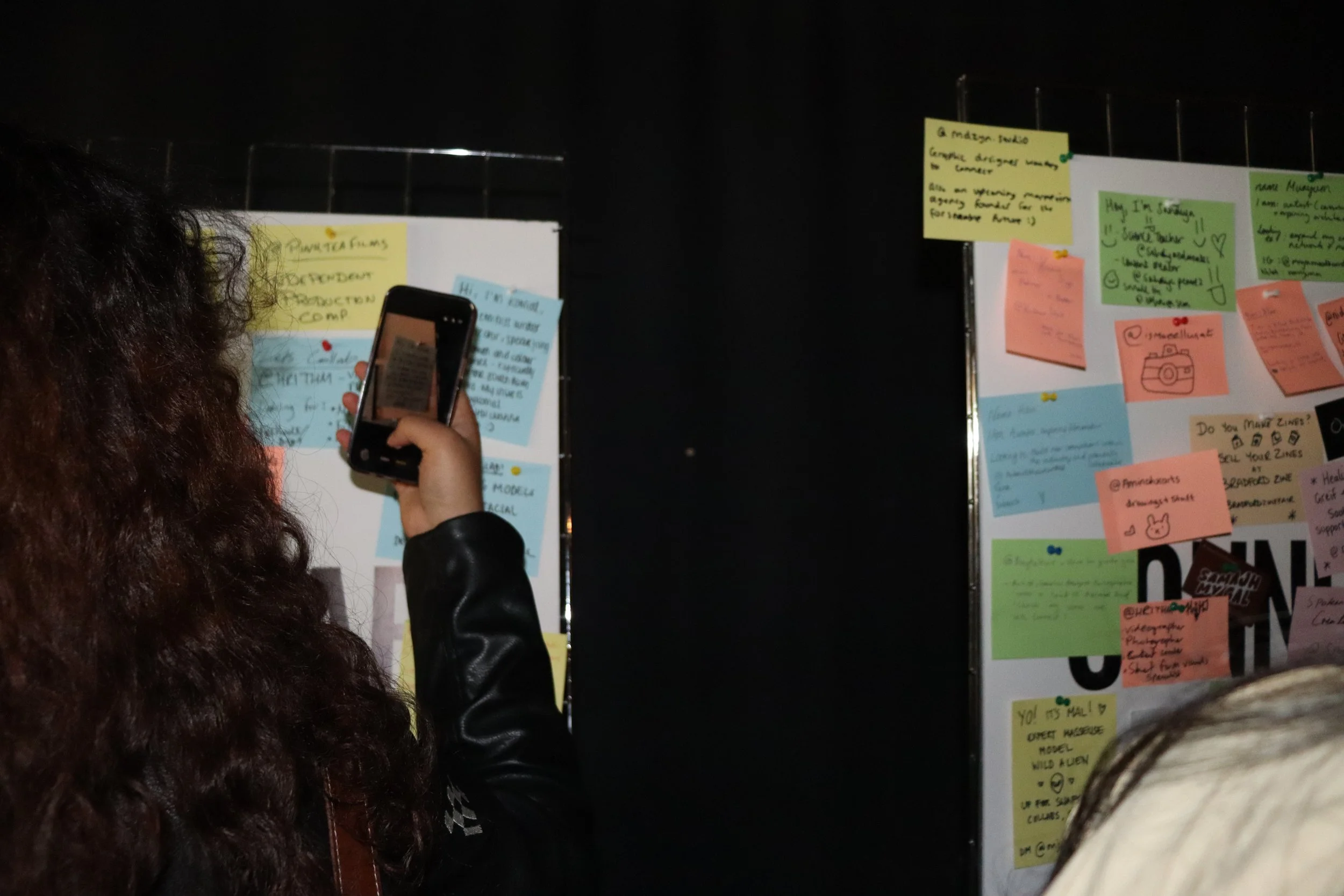 A person with curly hair taking a photo of a large bulletin board with their smartphone. The bulletin board is filled with colorful sticky notes and pinned papers containing handwritten notes and drawings.
