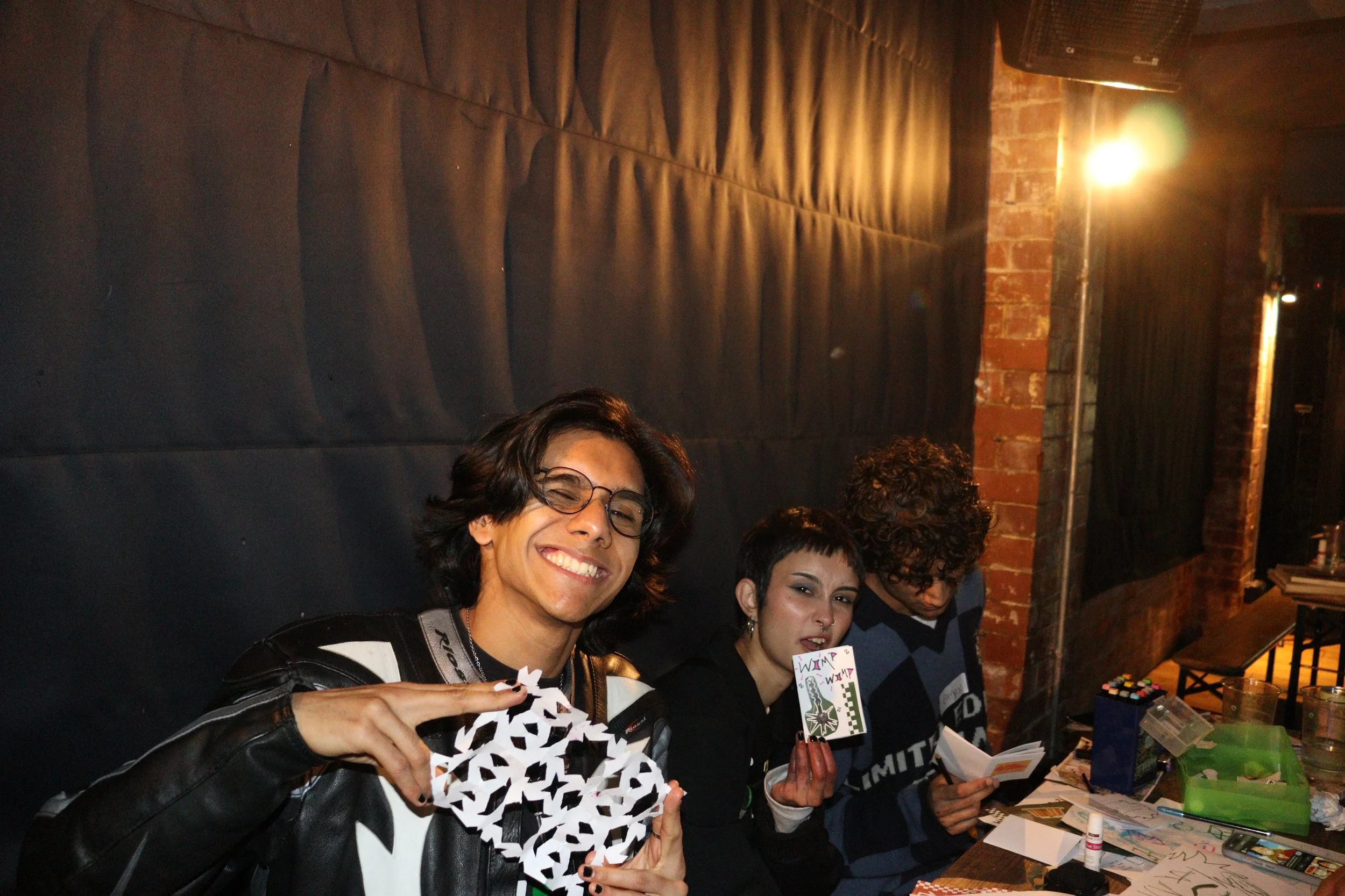 Group of four young people sitting at a table in an indoor setting, smiling and holding crafts and paper items, with a brick wall and warm lighting in the background.
