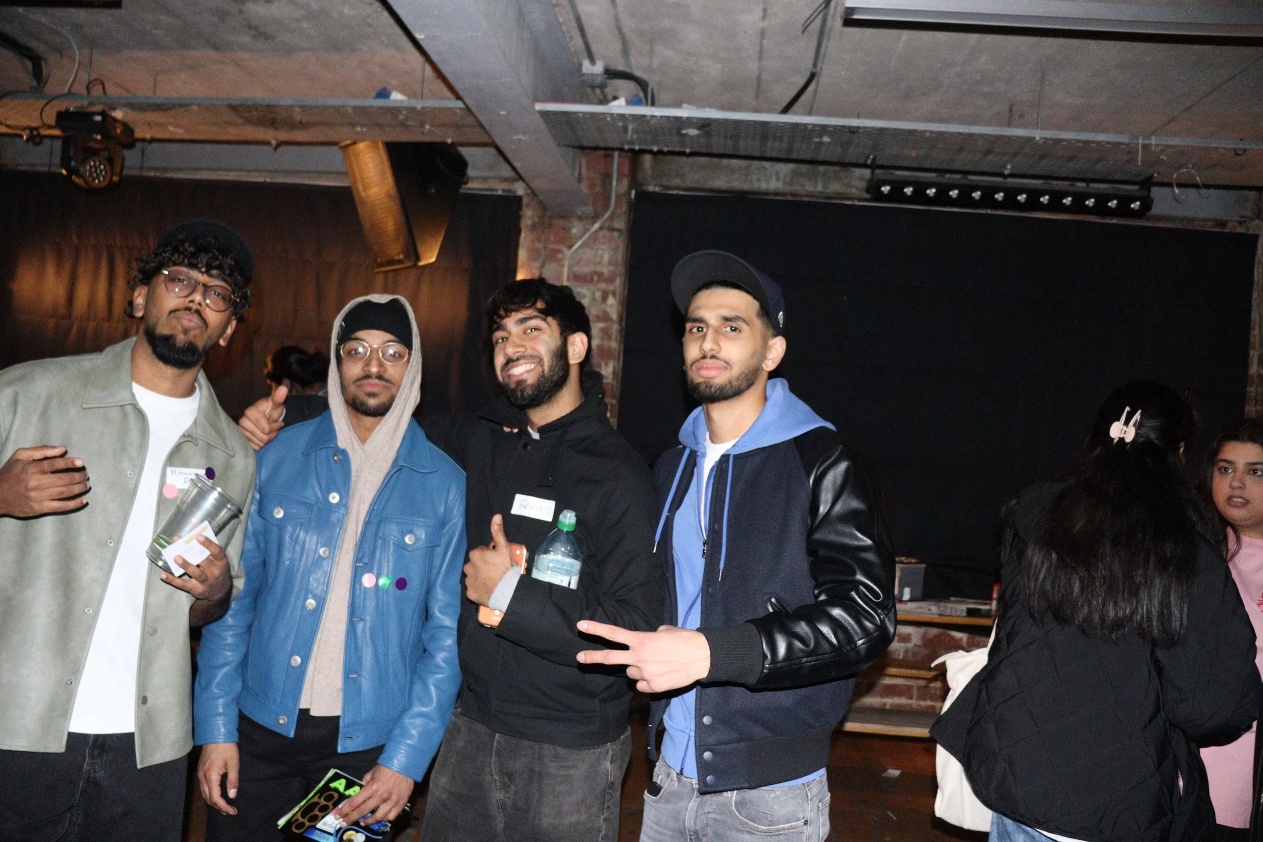 Four young men standing together at an indoor event, smiling and posing for the camera. They are wearing casual clothing, including jackets and hoodies, and one is holding a water bottle. The background shows a brick wall and some stage or event ligh