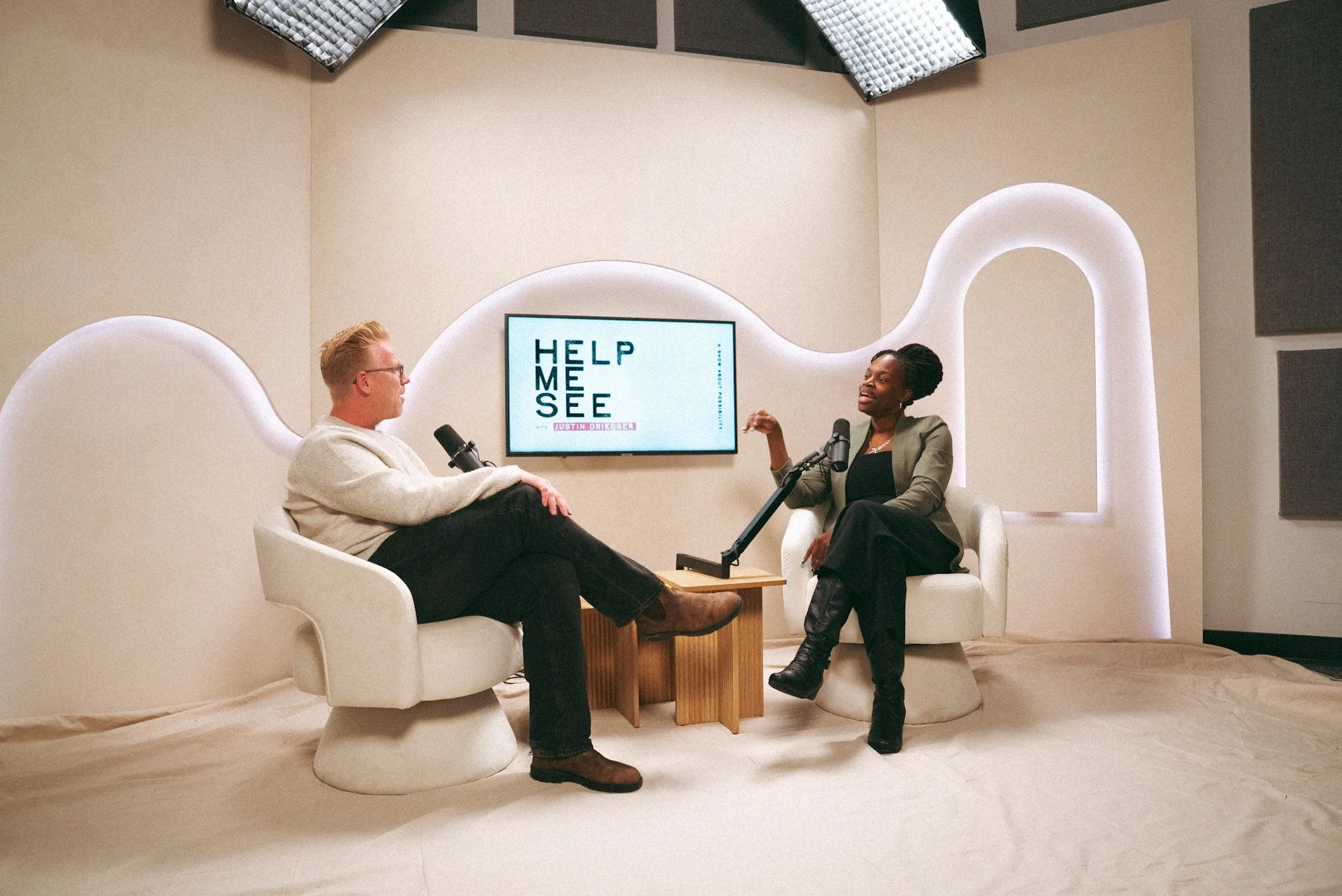 Two people sitting in a studio and having a conversation, with a sign on the wall behind them that says "Help Me See" with Justin Drieger's name.