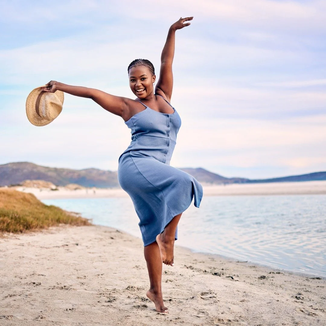 A woman dancing on a sandy beach near the water, holding a straw hat in her left hand, smiling, wearing a blue dress, during daytime with a partly cloudy sky.