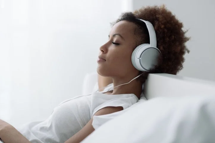 Young woman with natural hair wearing headphones and relaxing on a bed with white bedding.