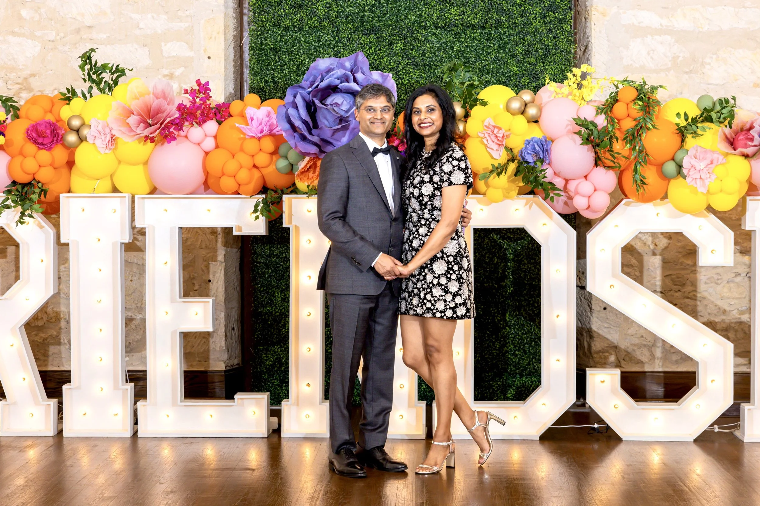 A couple stands in front of a decorative floral backdrop with large balloons and greenery, holding hands and smiling at a celebration or party.