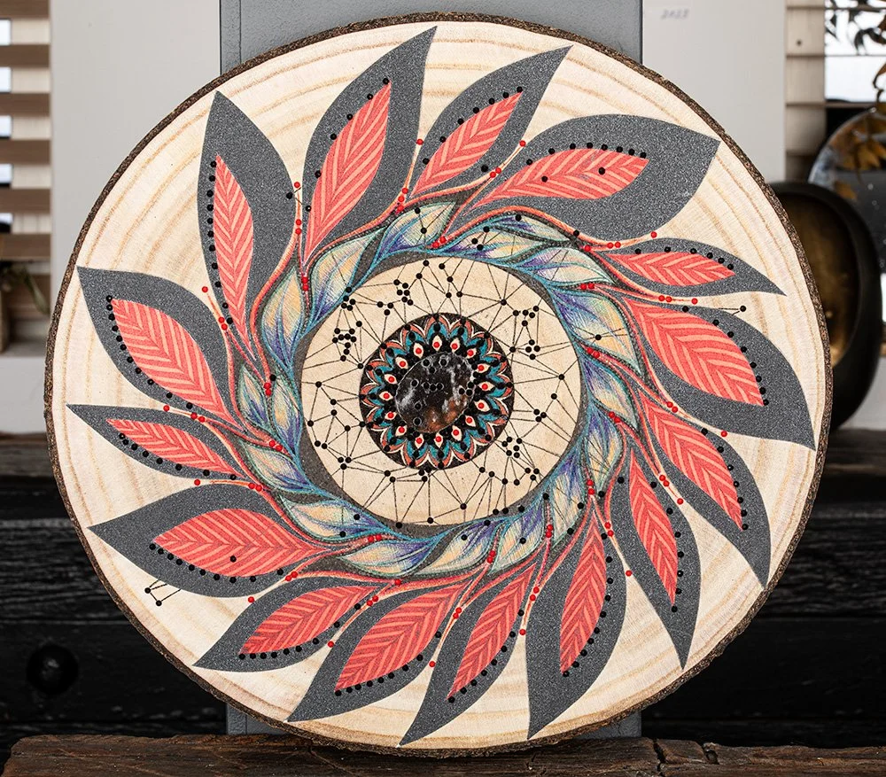 Mixed-media werk 'Indian Wheel' van Xandra Dupper: een spiraal van koraalrode en grijze bladeren rond een donkere steen in het midden, geschilderd op een ronde boomschijf.