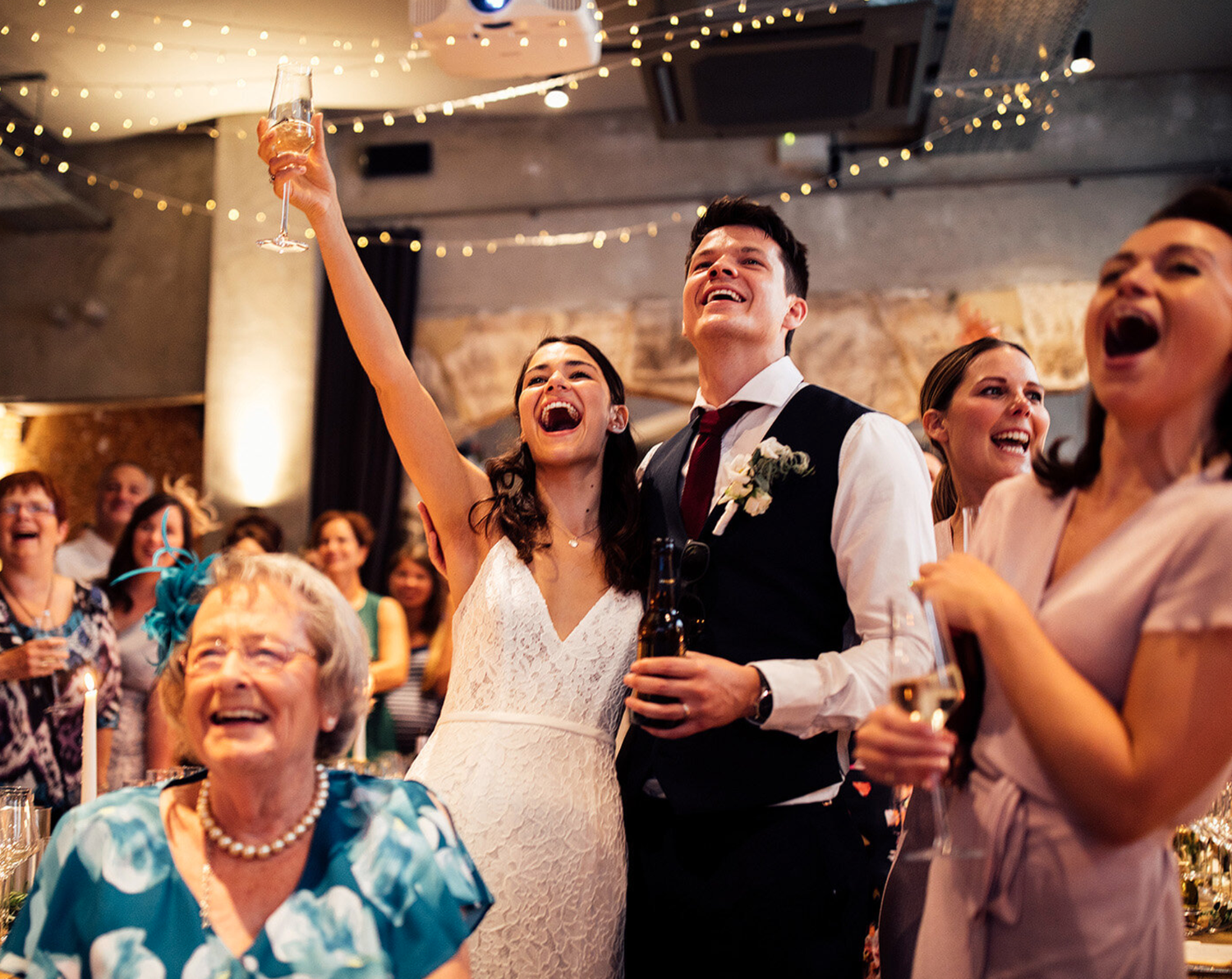 A wedding reception with a bride and groom smiling and laughing together at a decorated table with flowers, candles, and food, surrounded by guests.