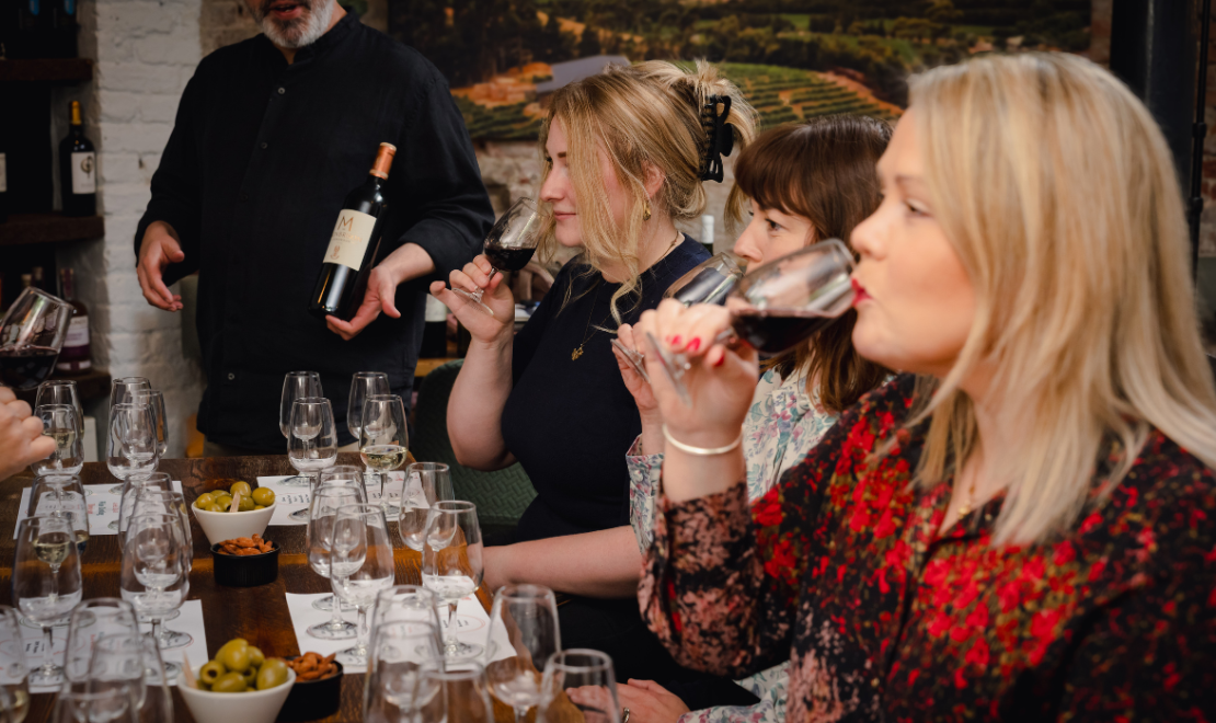 People tasting wine at a wine tasting event, with a waiter holding a bottle of red wine in the background.