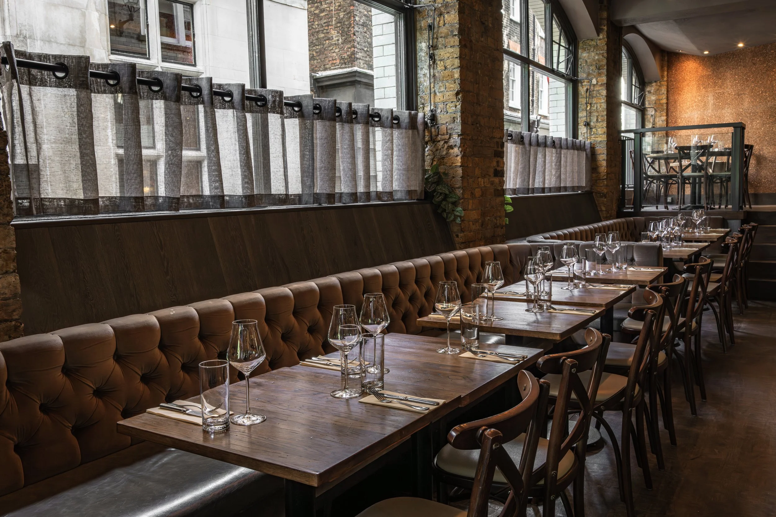 Interior of a restaurant with wooden tables, glasses set for dining, leather cushioned bench seating, large windows with surrounding brick walls, and a cozy ambiance.