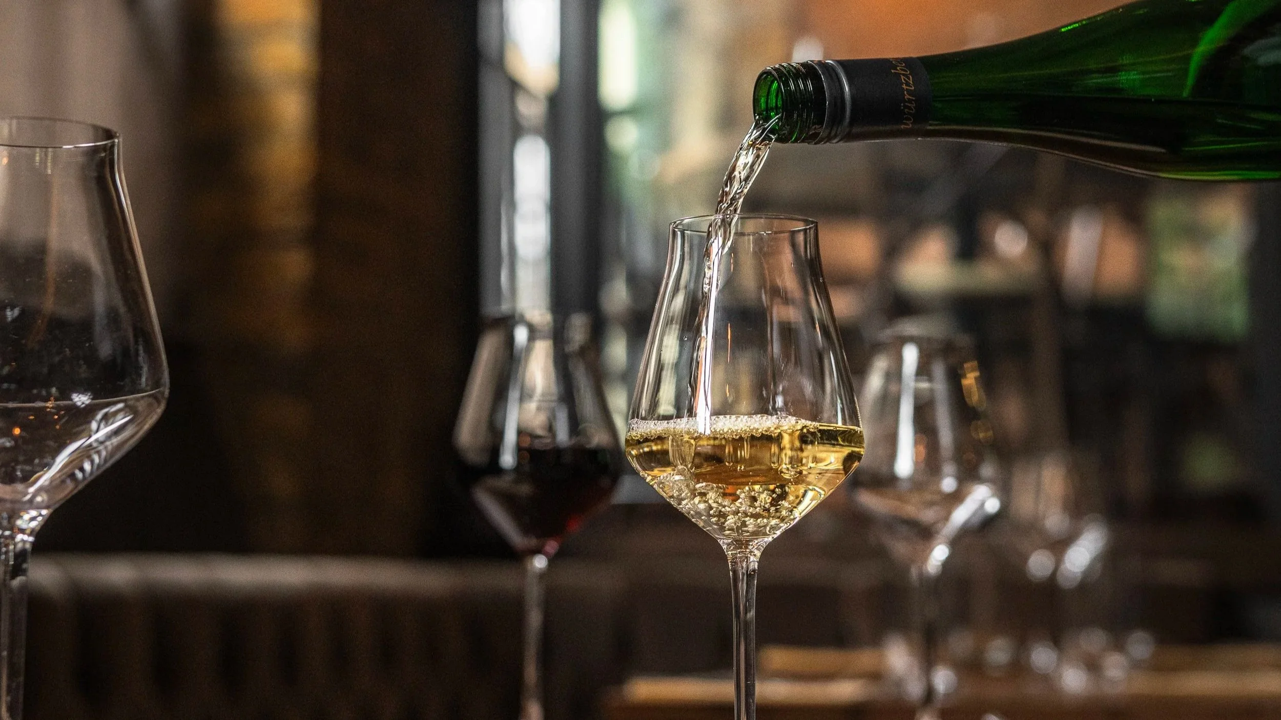 A bottle of white wine pouring into a wine glass on a bar or wine tasting table, with other glasses and bottles visible in the background.