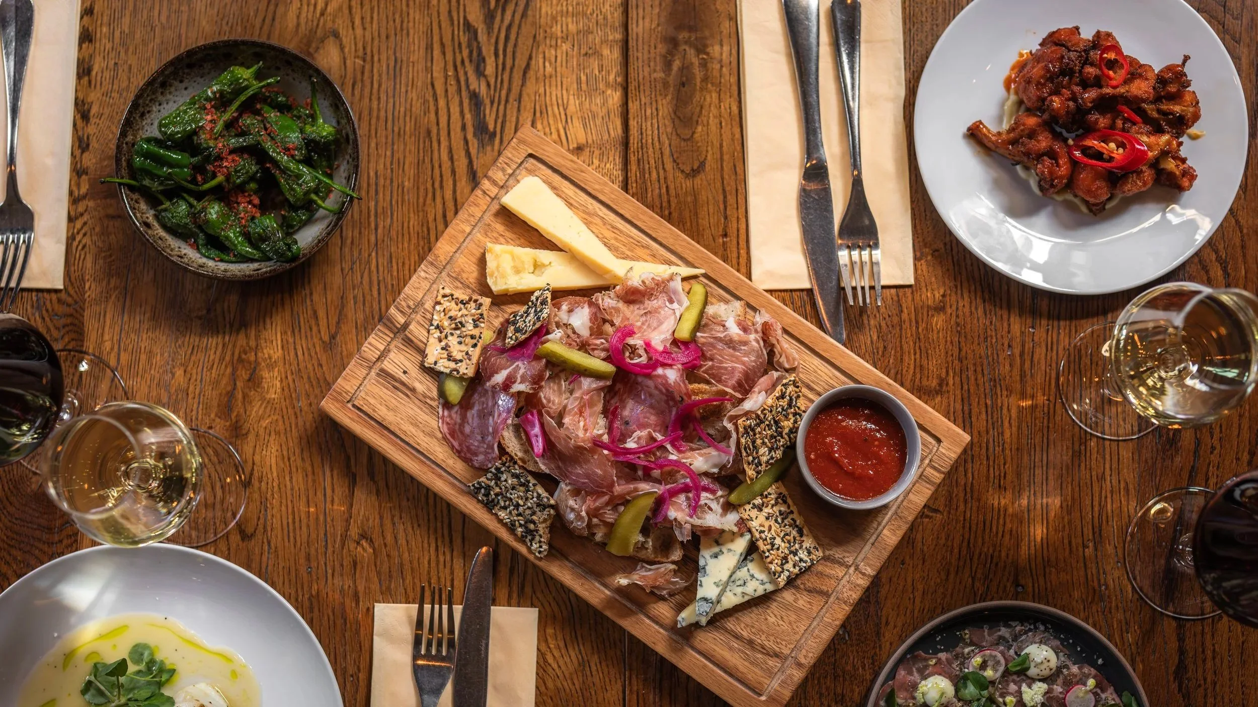 A wooden table with a cheese and charcuterie board, a bowl of greens, a plate of fried chicken with chili peppers, glasses of white wine, and bowls of soup or salad.