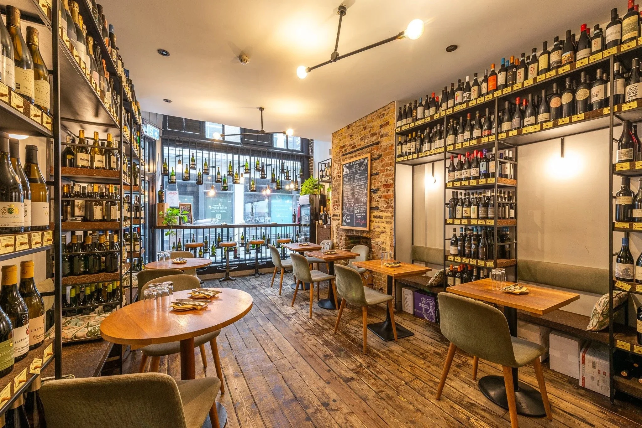 The Humble Grape Liverpool Street dining room, with wooden tables and a full wine wall beside exposed brick.
