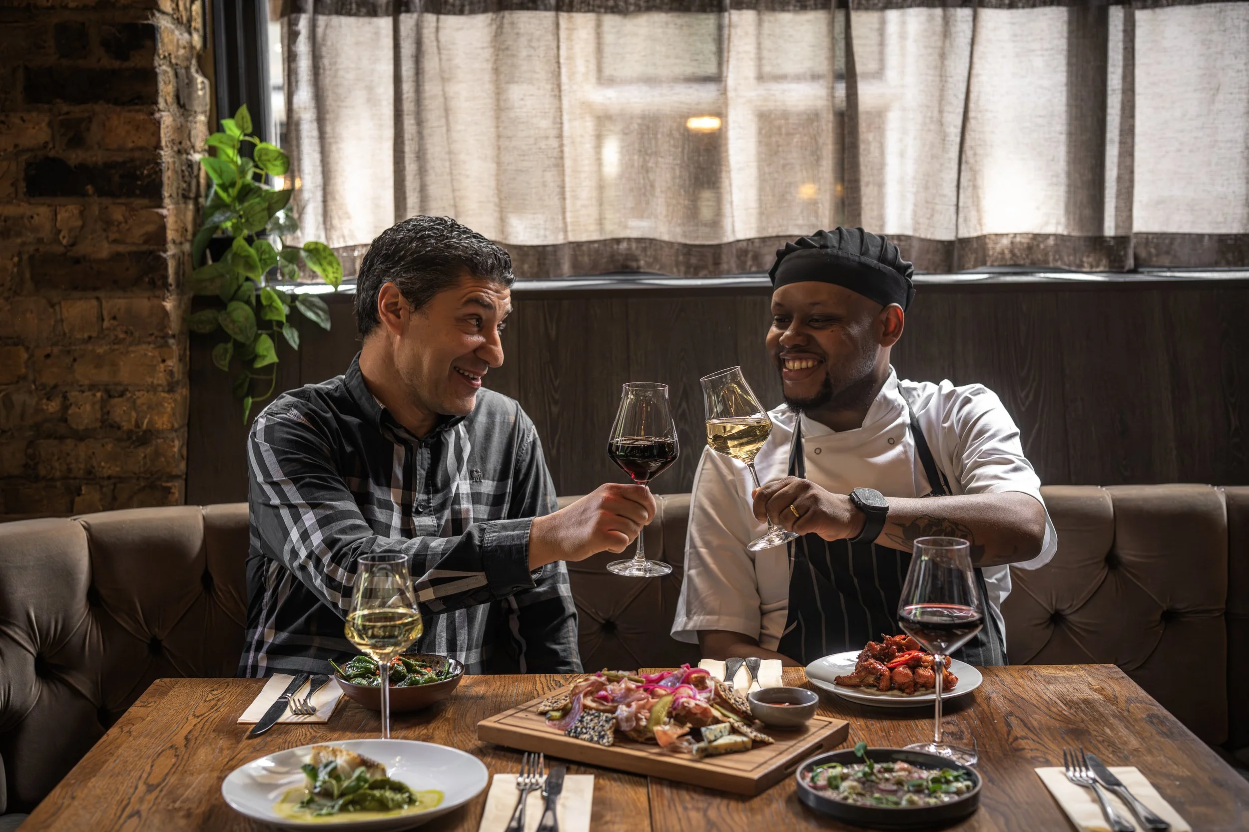 Two men are enjoying wine and dinner at a restaurant, smiling and toasting with glasses of white and red wine, with a spread of food on the table.