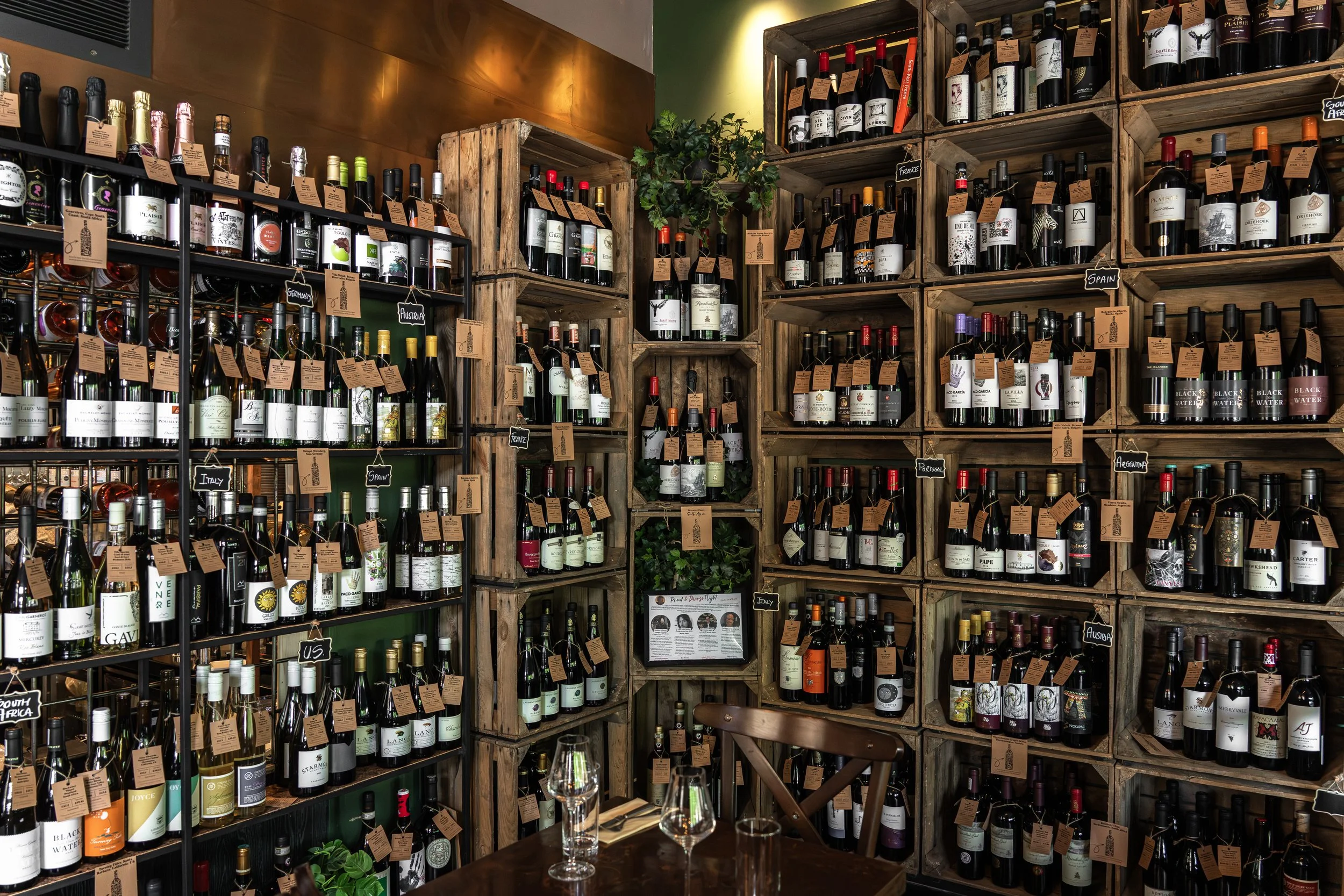 A wine shop with shelves filled with bottles of wine, each accompanied by a small brown tag. There is a small table in the foreground with two wine glasses and some green plants.